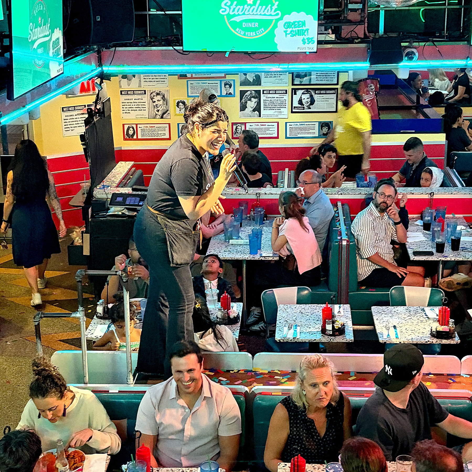 A lively diner is filled with customers sitting in booths, while a waitress joyfully interacts with guests from an elevated platform.