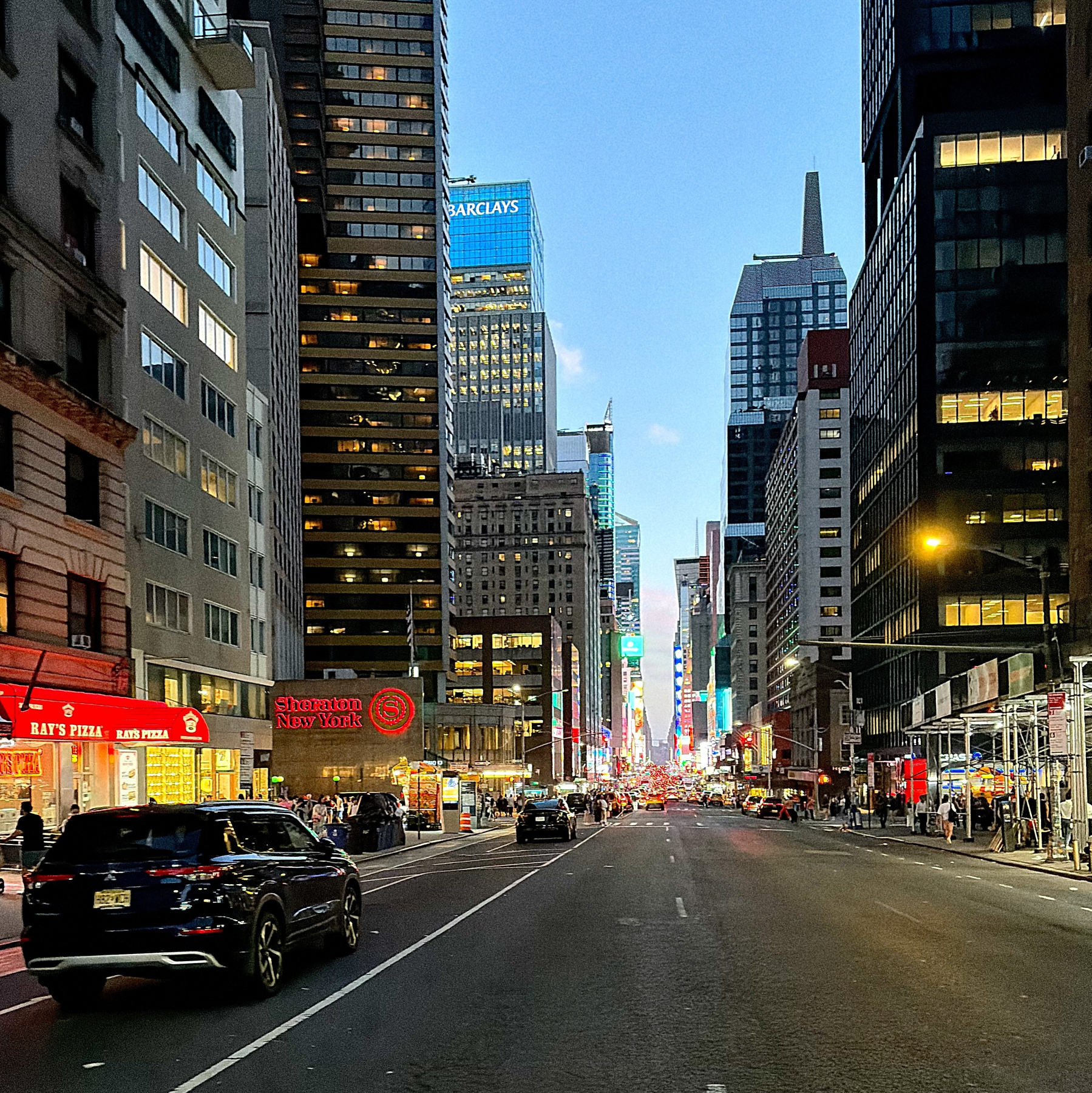 A busy city street at dusk is lined with tall buildings, illuminated signs, and cars on the road.