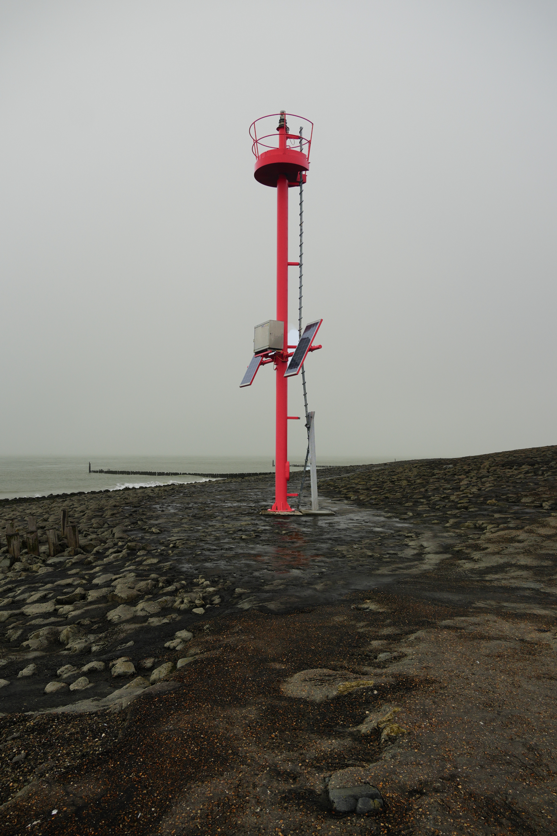 Auto-generated description: A red navigational beacon stands on a rocky shoreline under a cloudy sky.