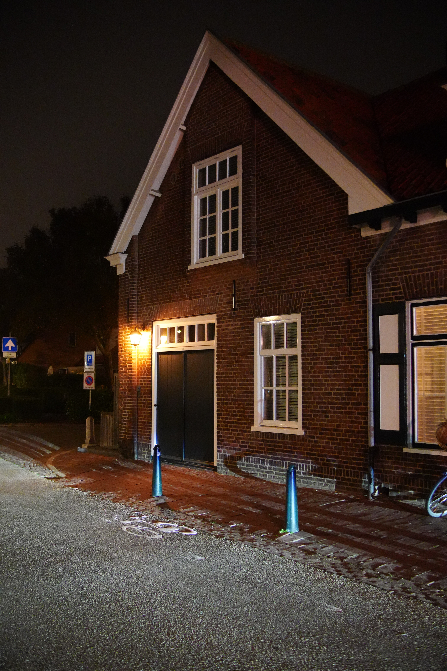 Auto-generated description: A brick building with a sharply peaked roof is illuminated by a warm streetlight at night, next to a road with visible cycling signs and blue bollards.