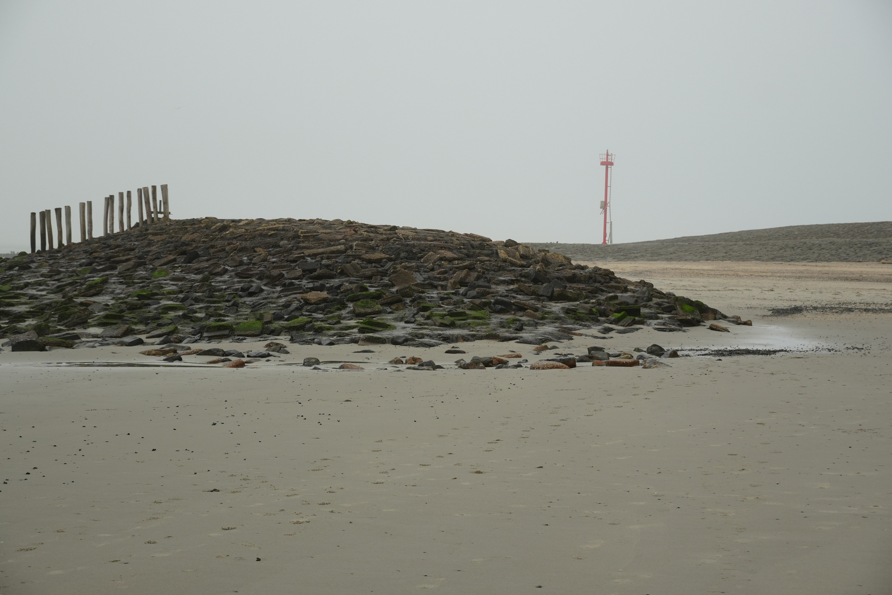 Auto-generated description: A rocky breakwater on a sandy beach with a distant red navigation marker.