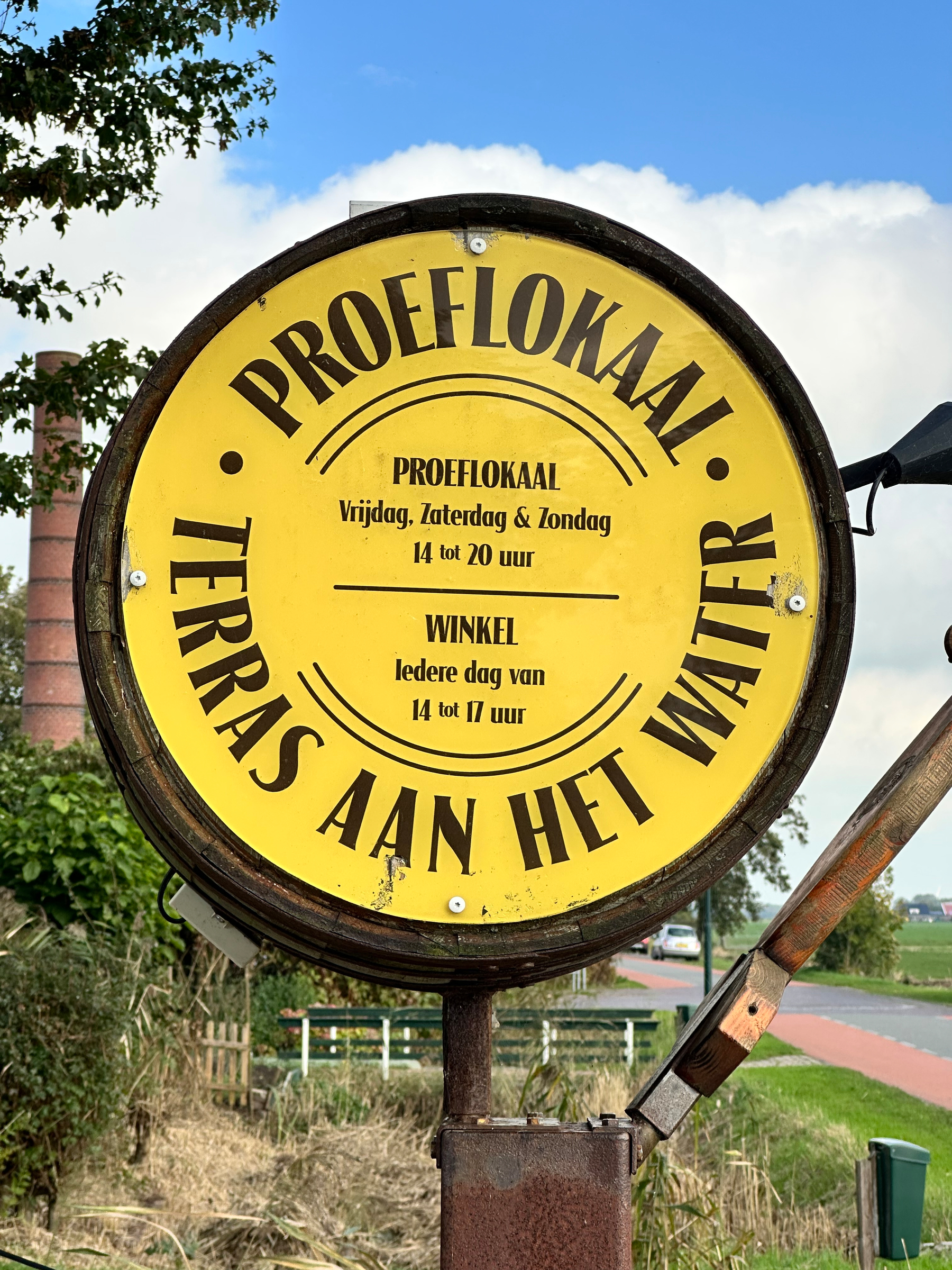 A yellow circular sign on a wooden barrel advertises a tasting room and waterside terrace with specific opening hours in Dutch.