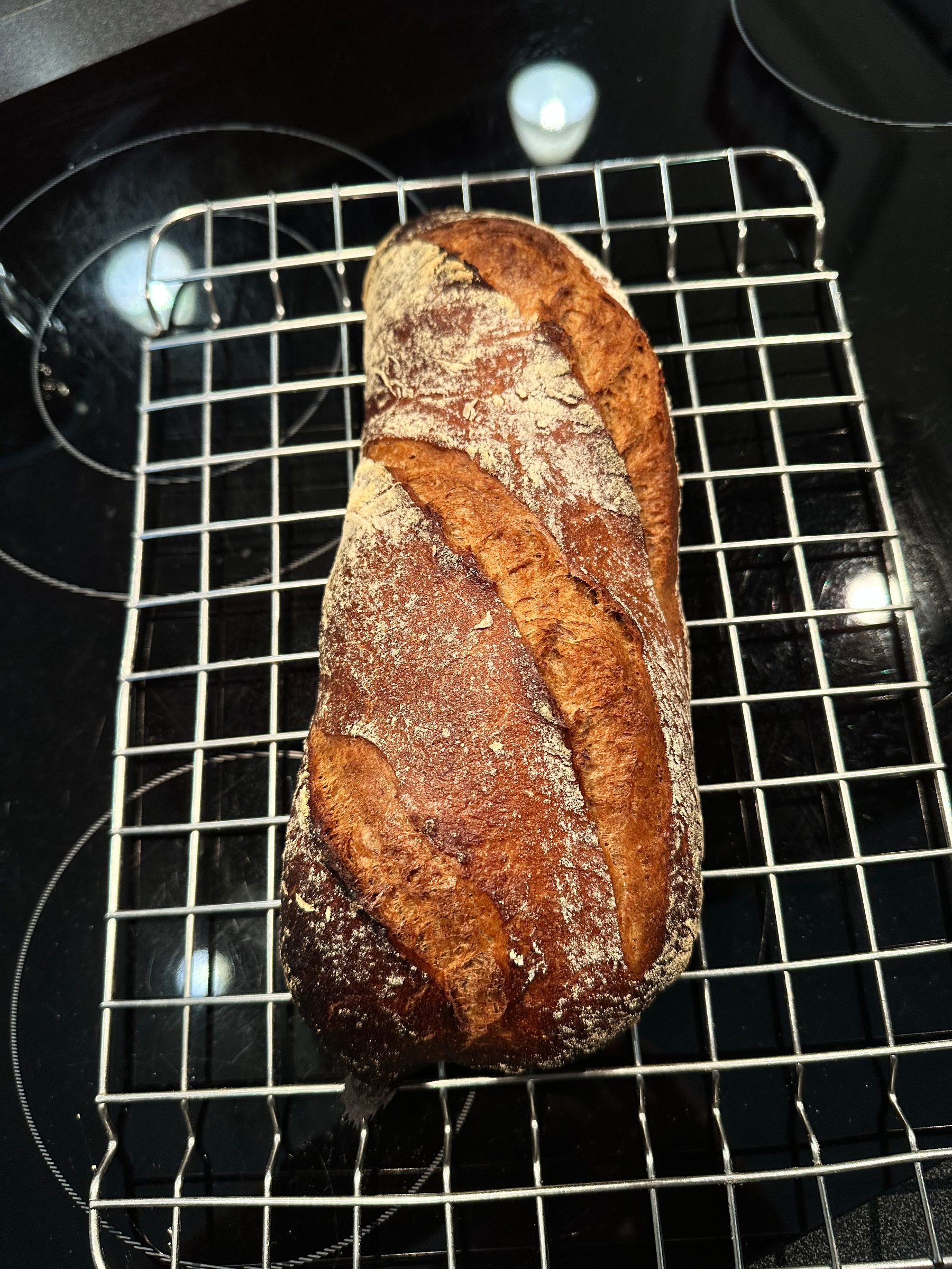 A freshly baked loaf of rustic bread is cooling on a wire rack placed on a black stovetop.