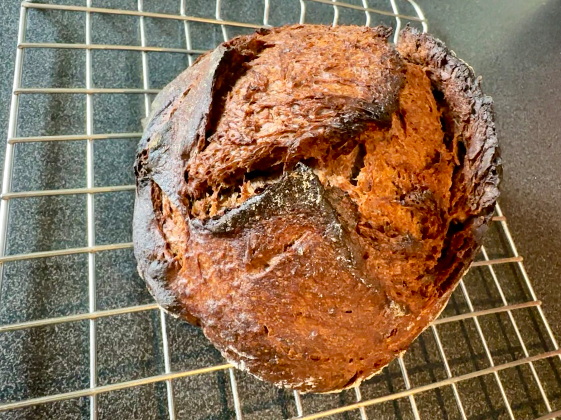 A round loaf of bread with a dark, crispy crust is resting on a wire cooling rack.