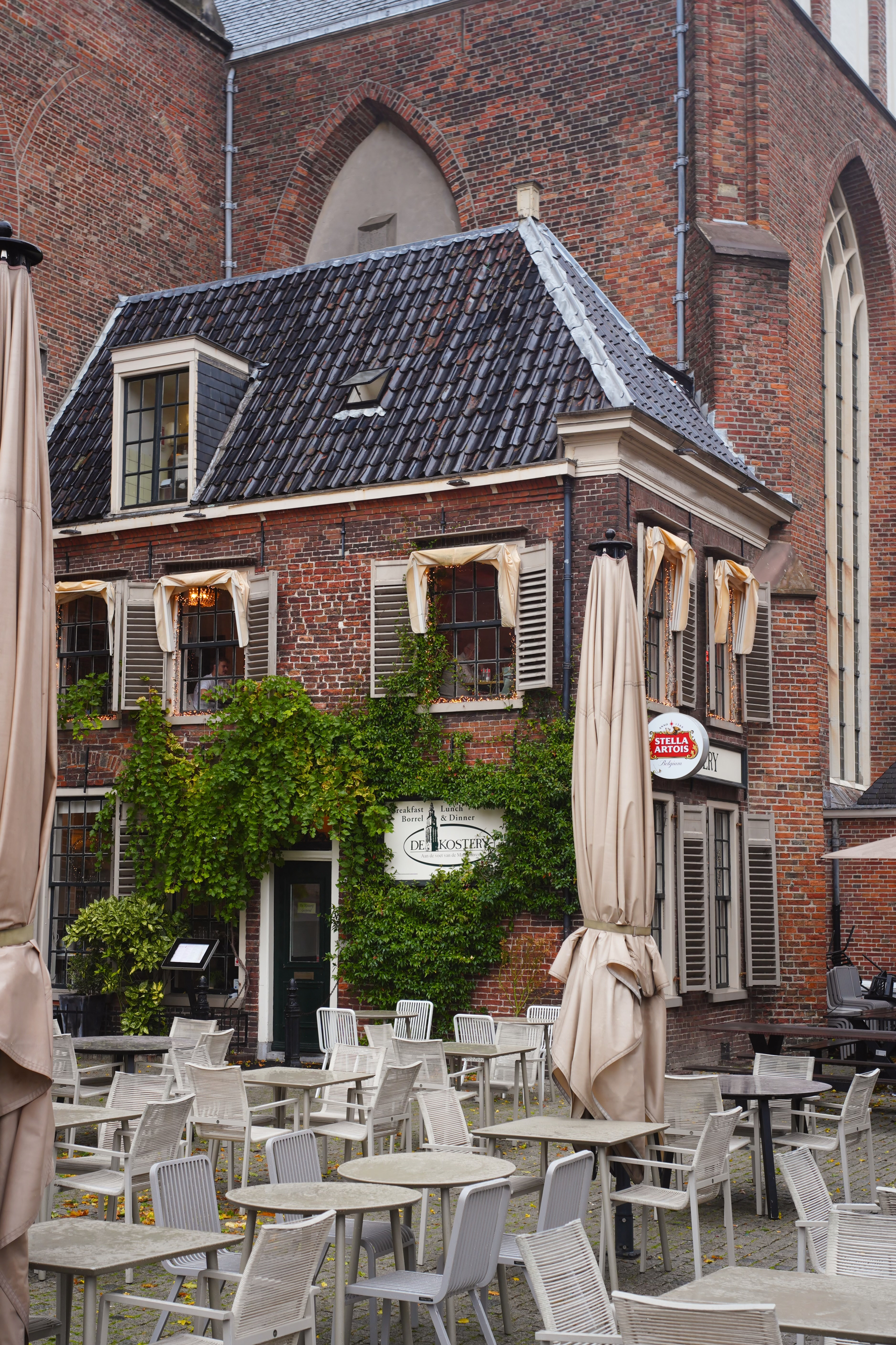 A quaint, brick-walled café with outdoor seating and green ivy growing on the walls sits nestled against a larger brick building.