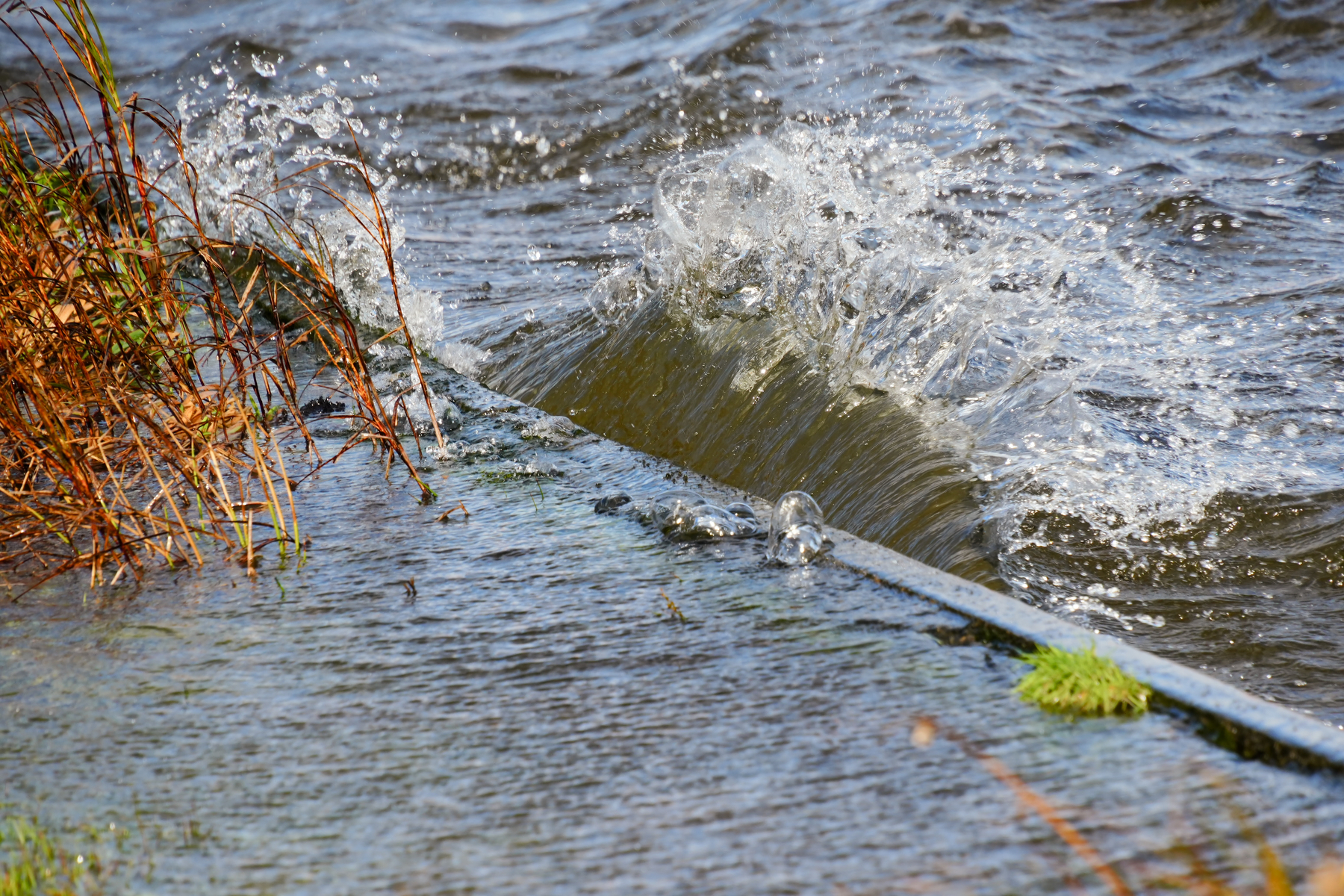 Waves are splashing over a metal or concrete edge along a water body with some grass and plants growing near the edge.