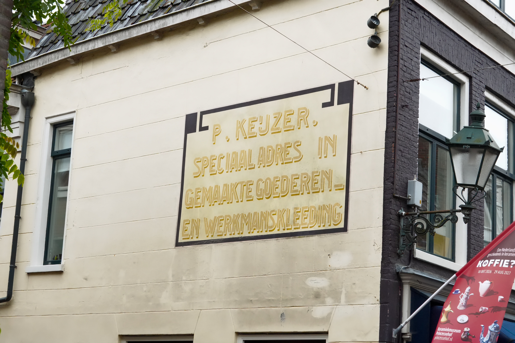 A beige wall features a vintage sign with Dutch text advertising P. Keuzer, special address in made goods and work clothing, next to a dark-trimmed building corner with a lamp and part of a red umbrella.