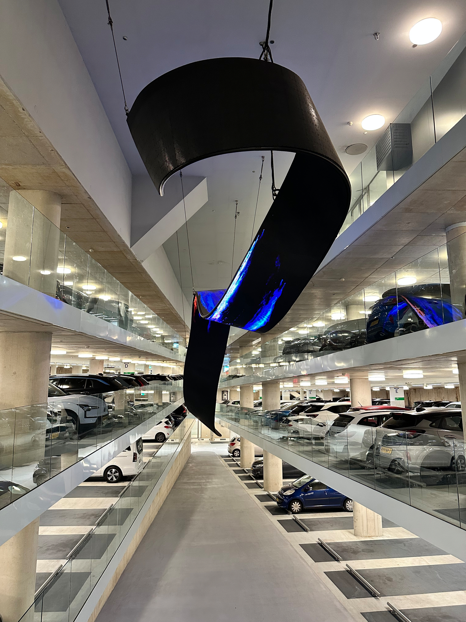 A modern multi-level parking garage features a large, curved black sculpture suspended from the ceiling above an empty driving lane.