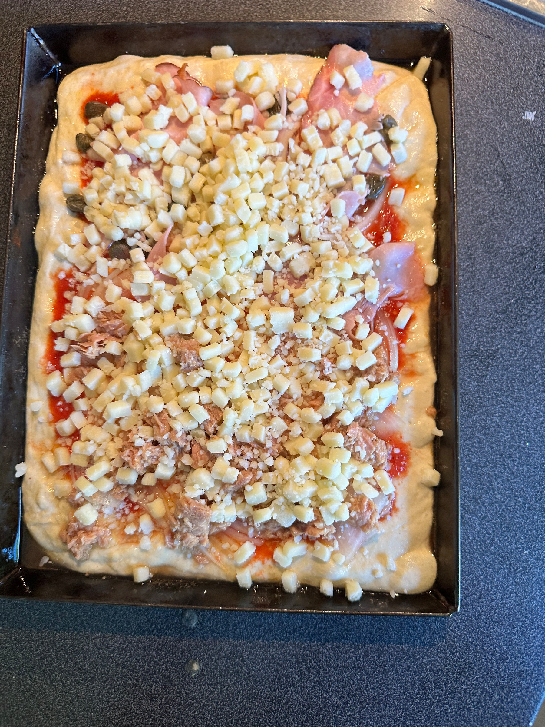 A rectangular pizza dough in a black baking tray is topped with tomato sauce, ham, tuna, capers, and shredded cheese.