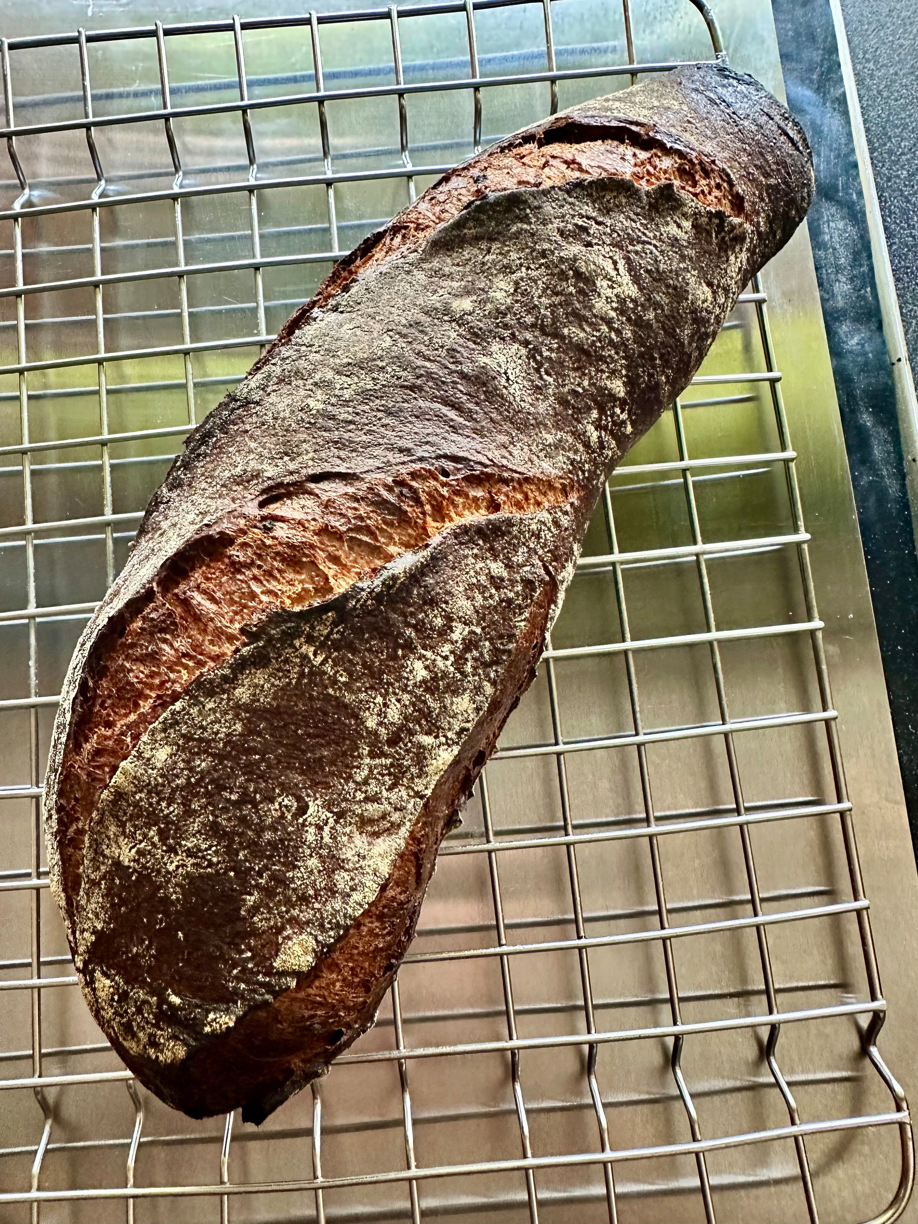A rustic loaf of bread with a dark crust rests on a cooling rack.