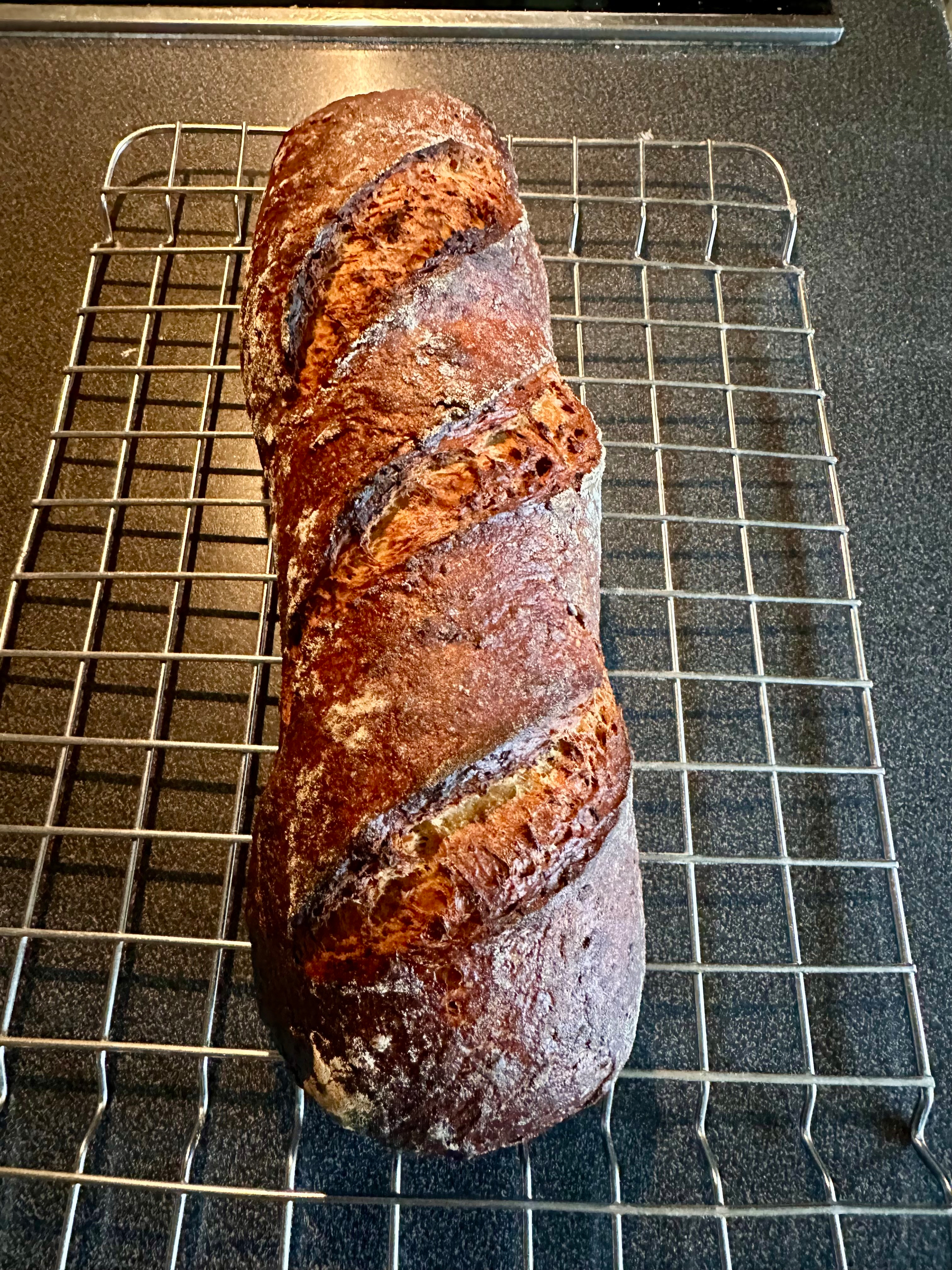 A loaf of crusty, artisan bread with deep slashes on top is cooling on a wire rack.