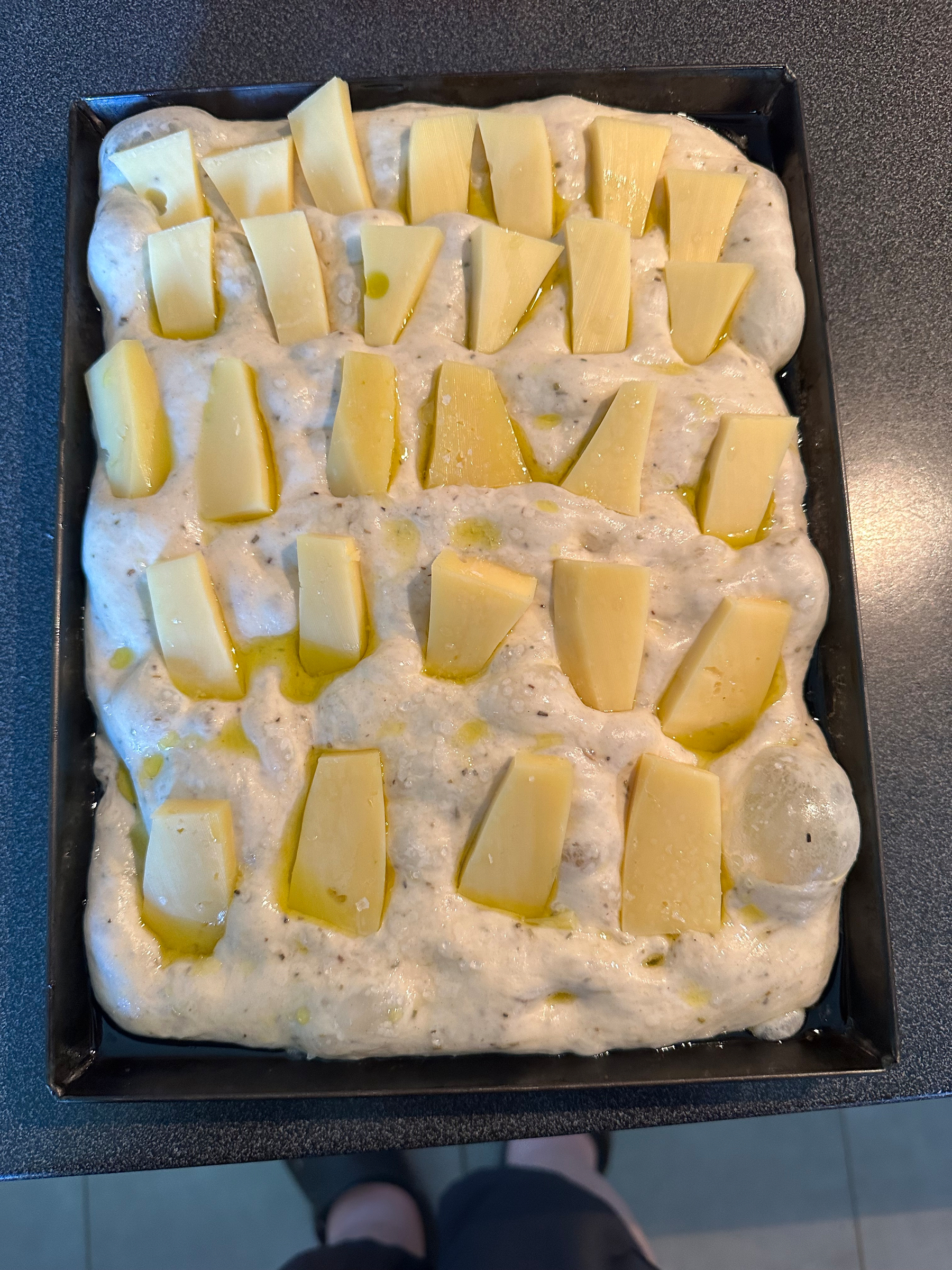 A focaccia dough is spread out in a rectangular baking pan with pieces of cheese and drizzled olive oil on top.