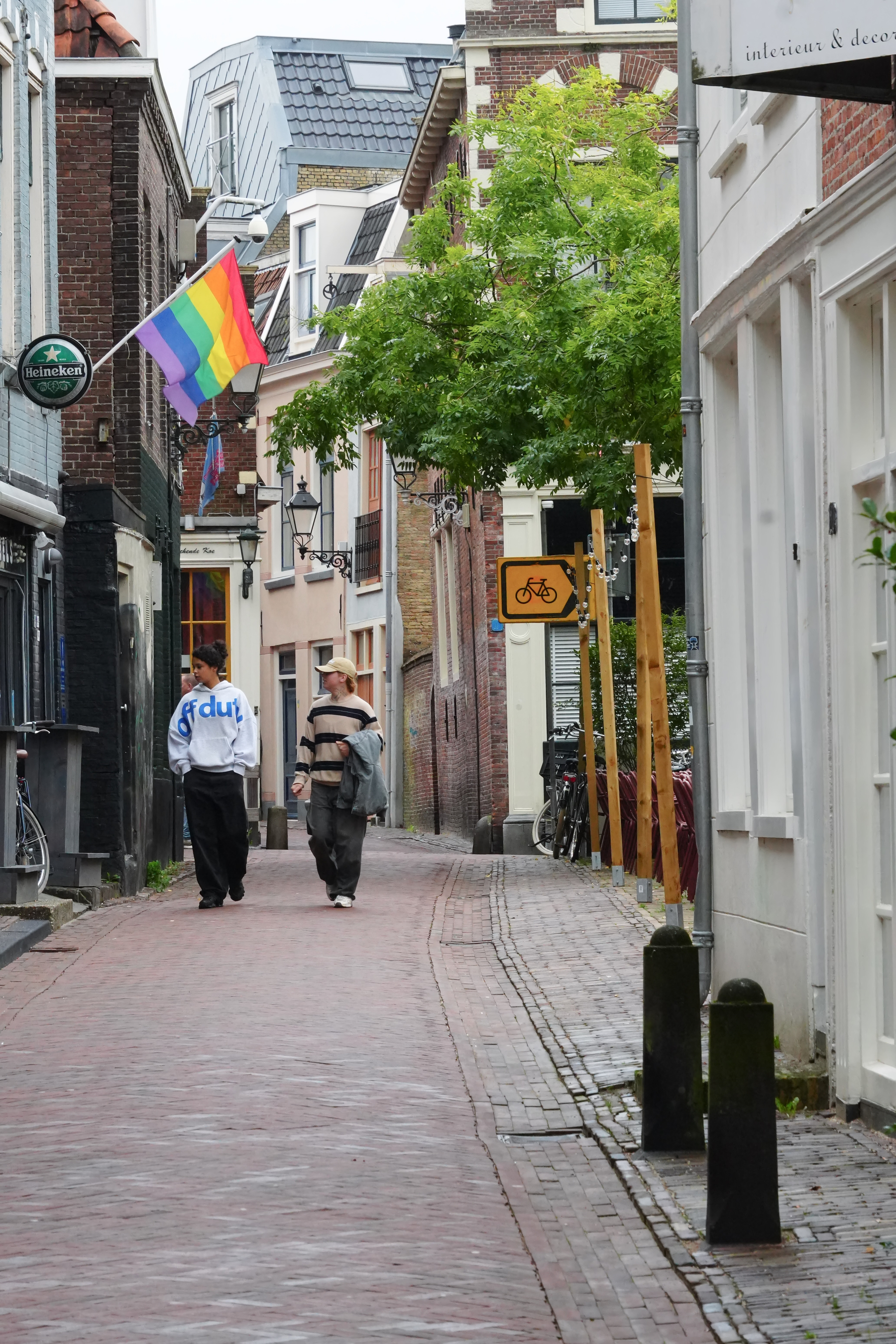 Two people are walking down a narrow cobblestone street lined with buildings, a rainbow flag, and a bicycle sign visible.
