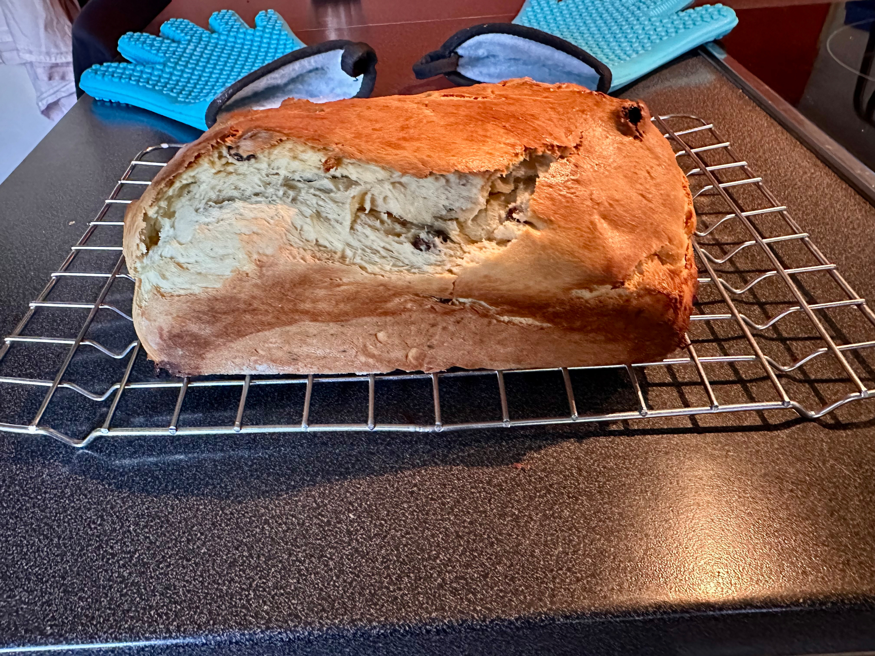 A loaf of brioche is cooling on a wire rack with a pair of blue oven mitts in the background.
