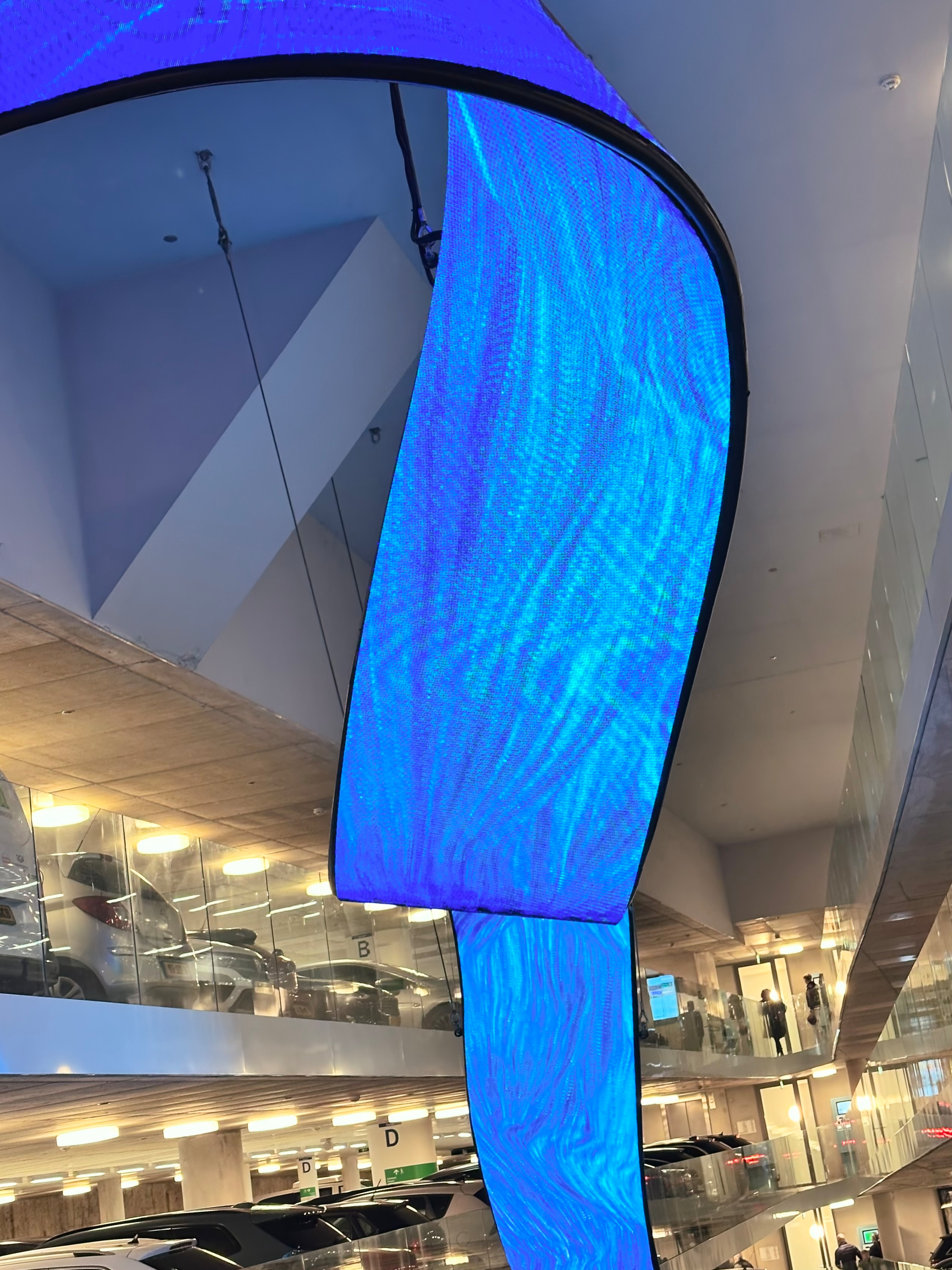 A large, curved digital screen displaying shades of blue is suspended in a multi-level indoor parking garage.