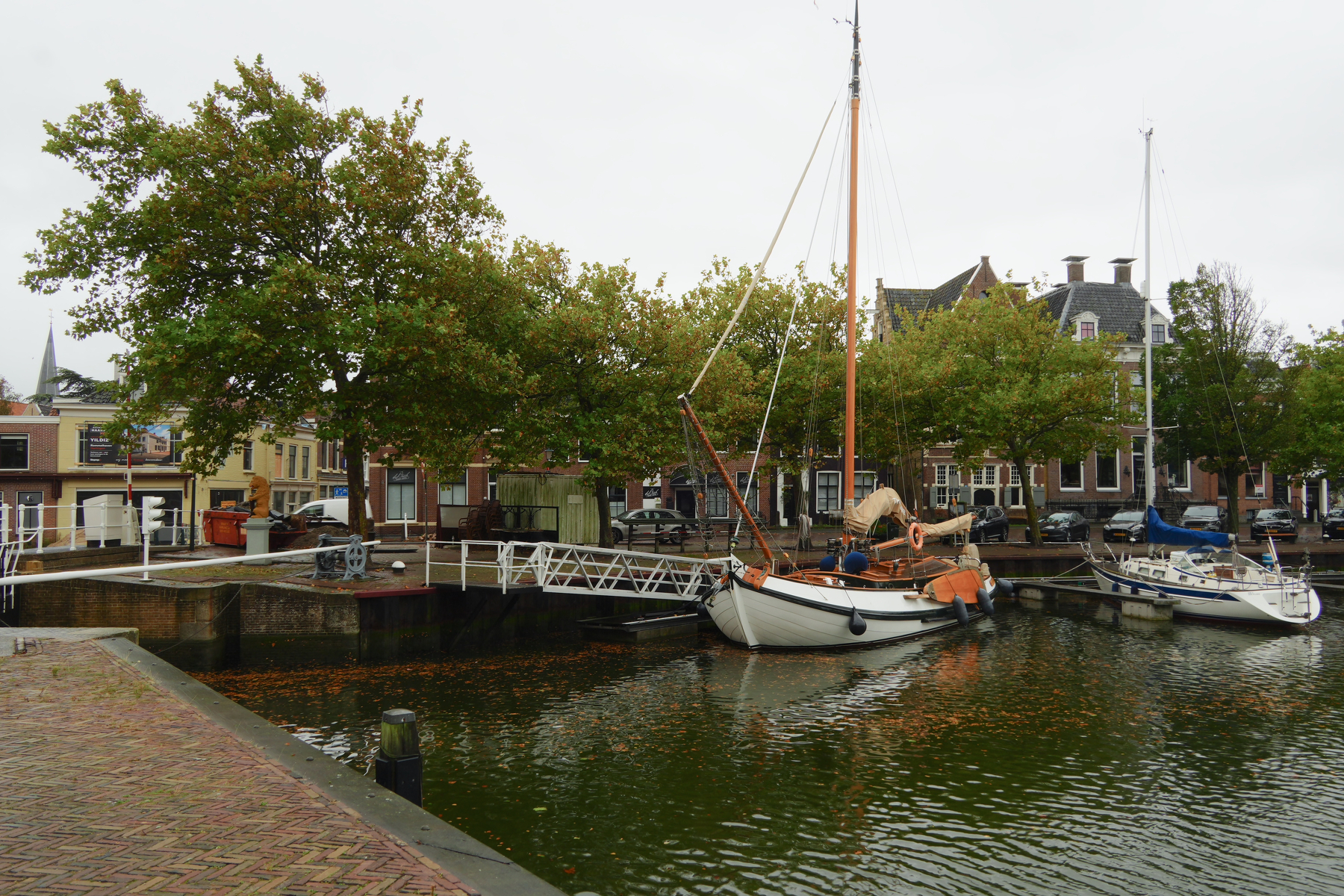 A small harbor with several sailboats docked alongside a walkway lined with trees and buildings in the background.