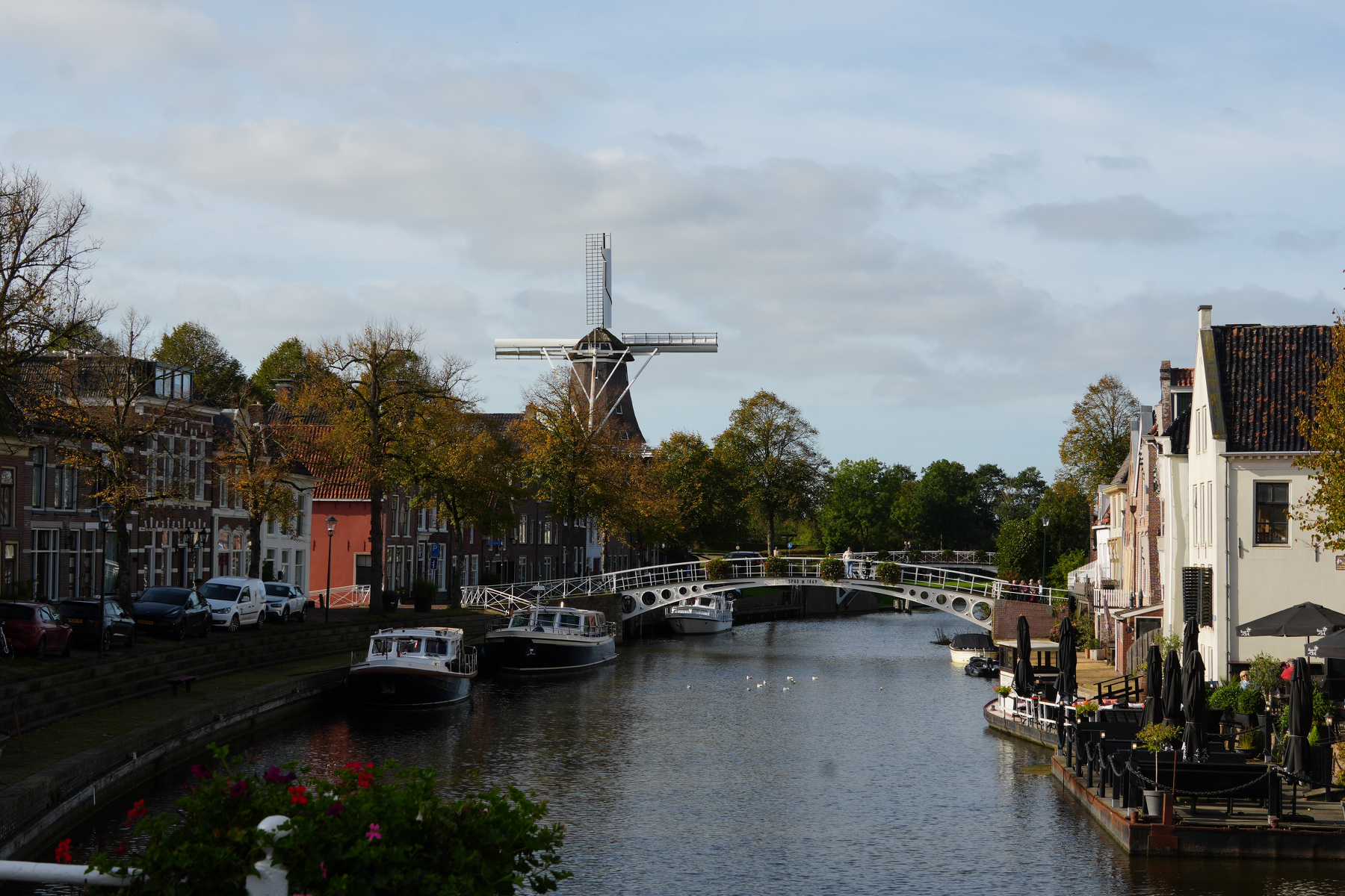 A peaceful canal is lined with boats and traditional buildings, with a windmill and a bridge in the background under a partly cloudy sky. Dokkum