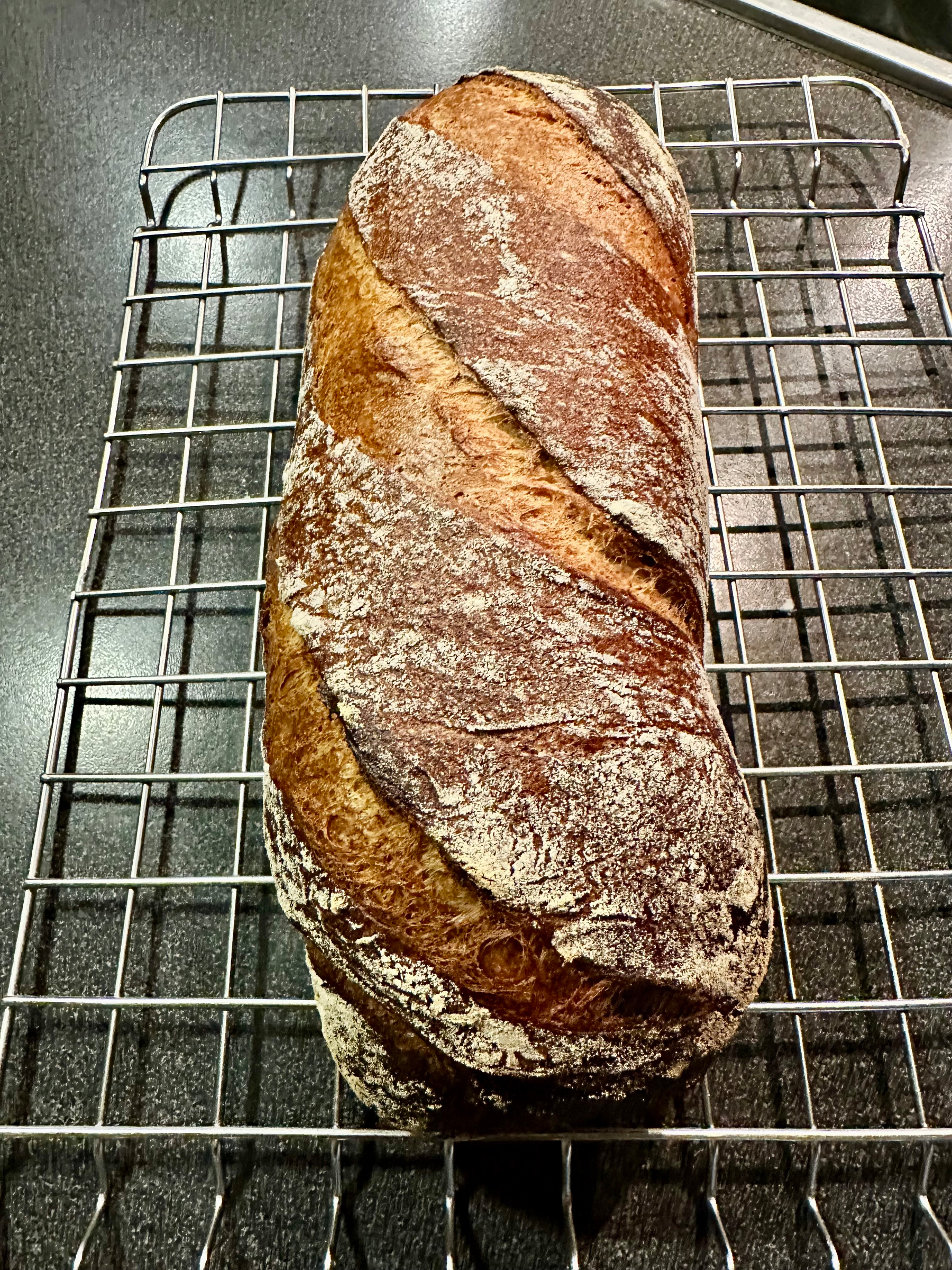 A freshly baked loaf of rustic bread with a golden-brown crust is cooling on a metal rack.
