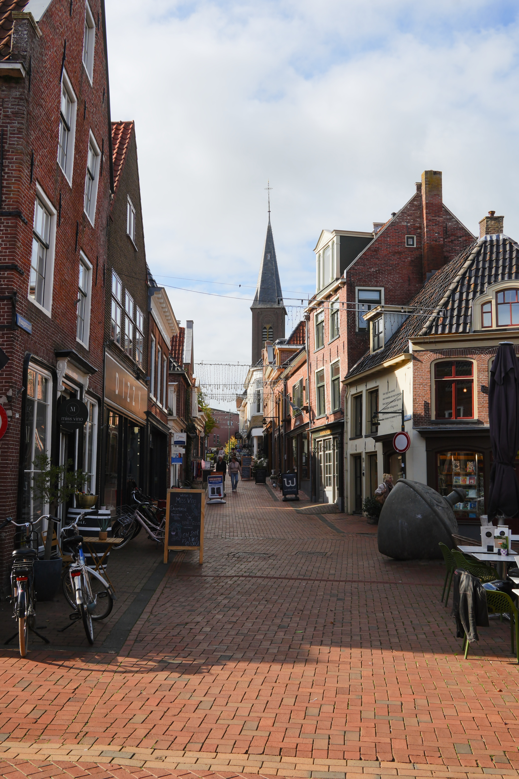 A narrow cobblestone street lined with brick buildings, bicycles, outdoor seating, and a church steeple in the background is visible.