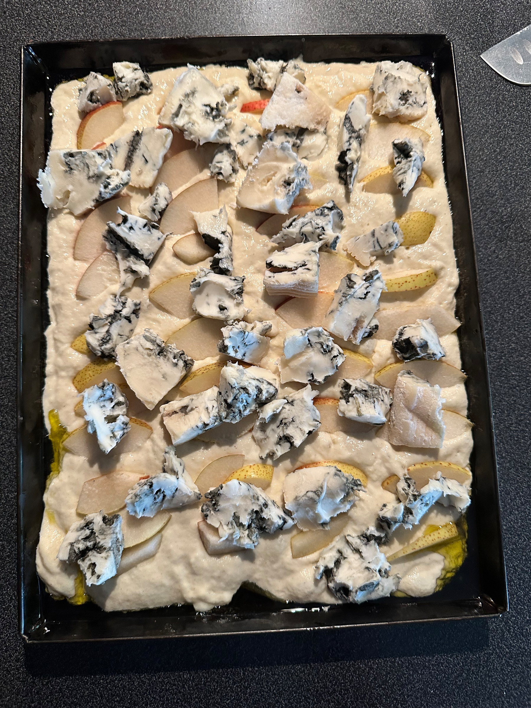 A rectangular baking dish contains focaccia  dough topped with slices of pear and chunks of blue-veined cheese.