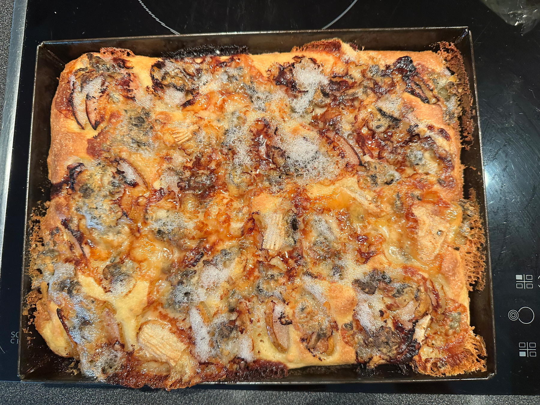 A rectangular homemade focaccia with pears and melted Gorgonzola is cooling in a black baking tray on a stovetop.