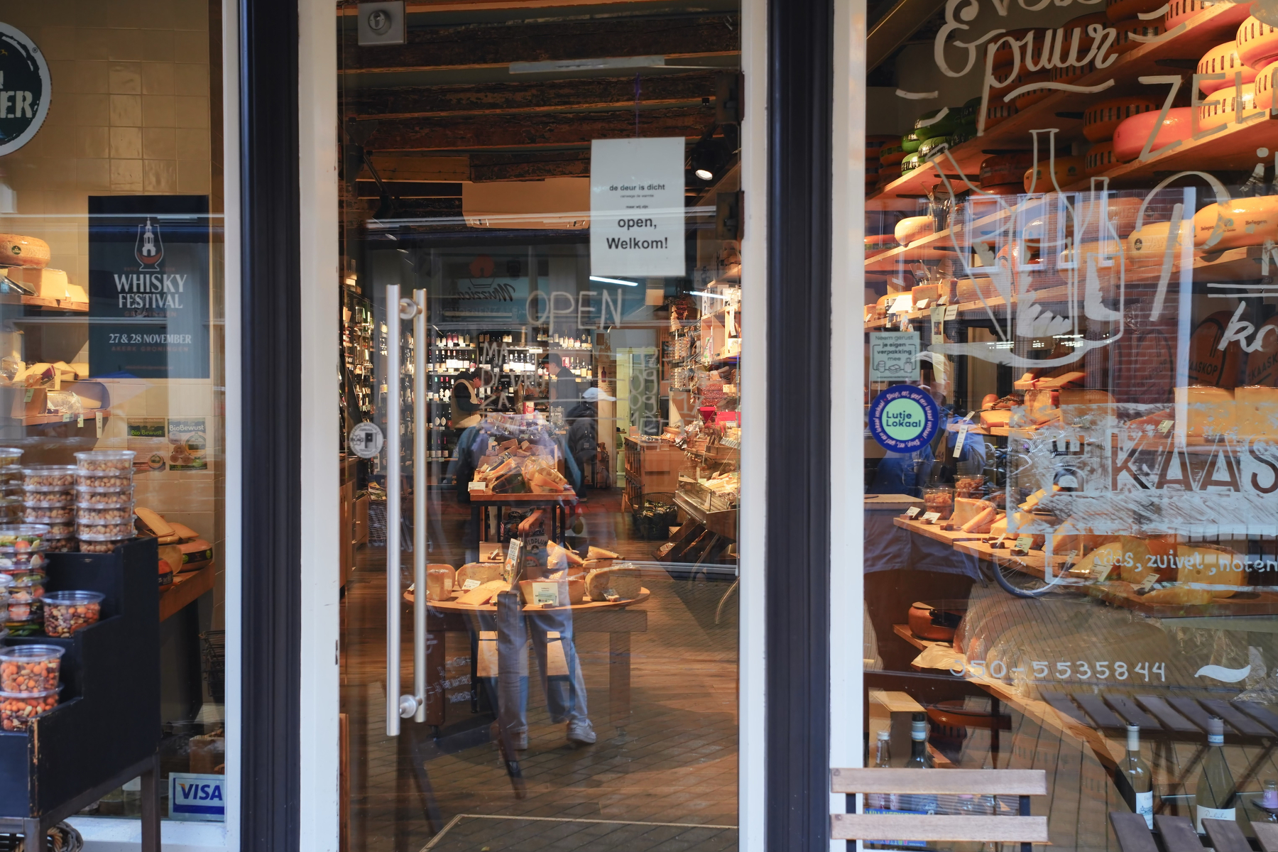 A warmly lit shop interior displays a variety of bread and baked goods with a sign on the door saying open, welkom! in Dutch.