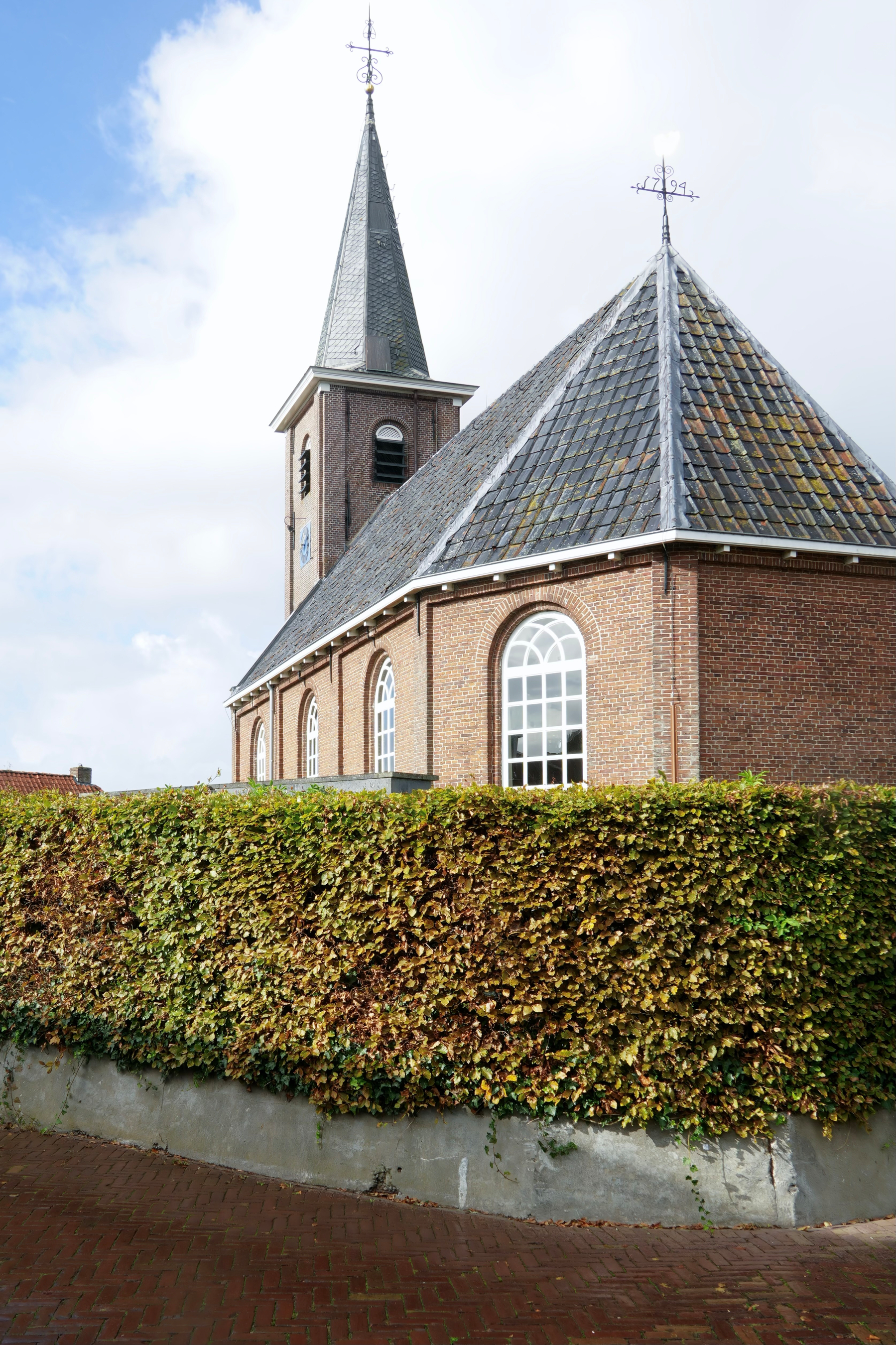 A brick church with a tall steeple and arched windows is partially obscured by a neatly trimmed hedge under a partly cloudy sky.