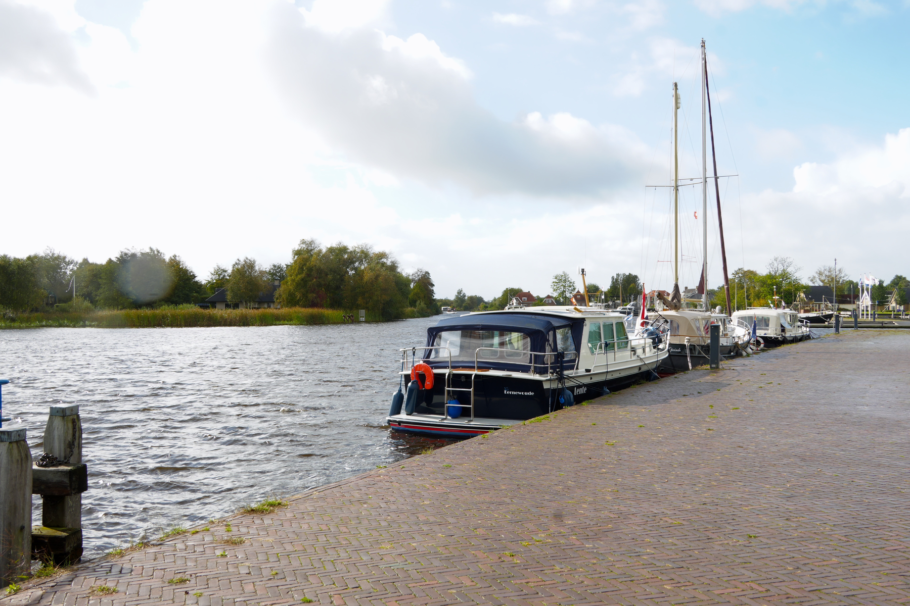 Several boats are docked along a paved riverbank under a partly cloudy sky.