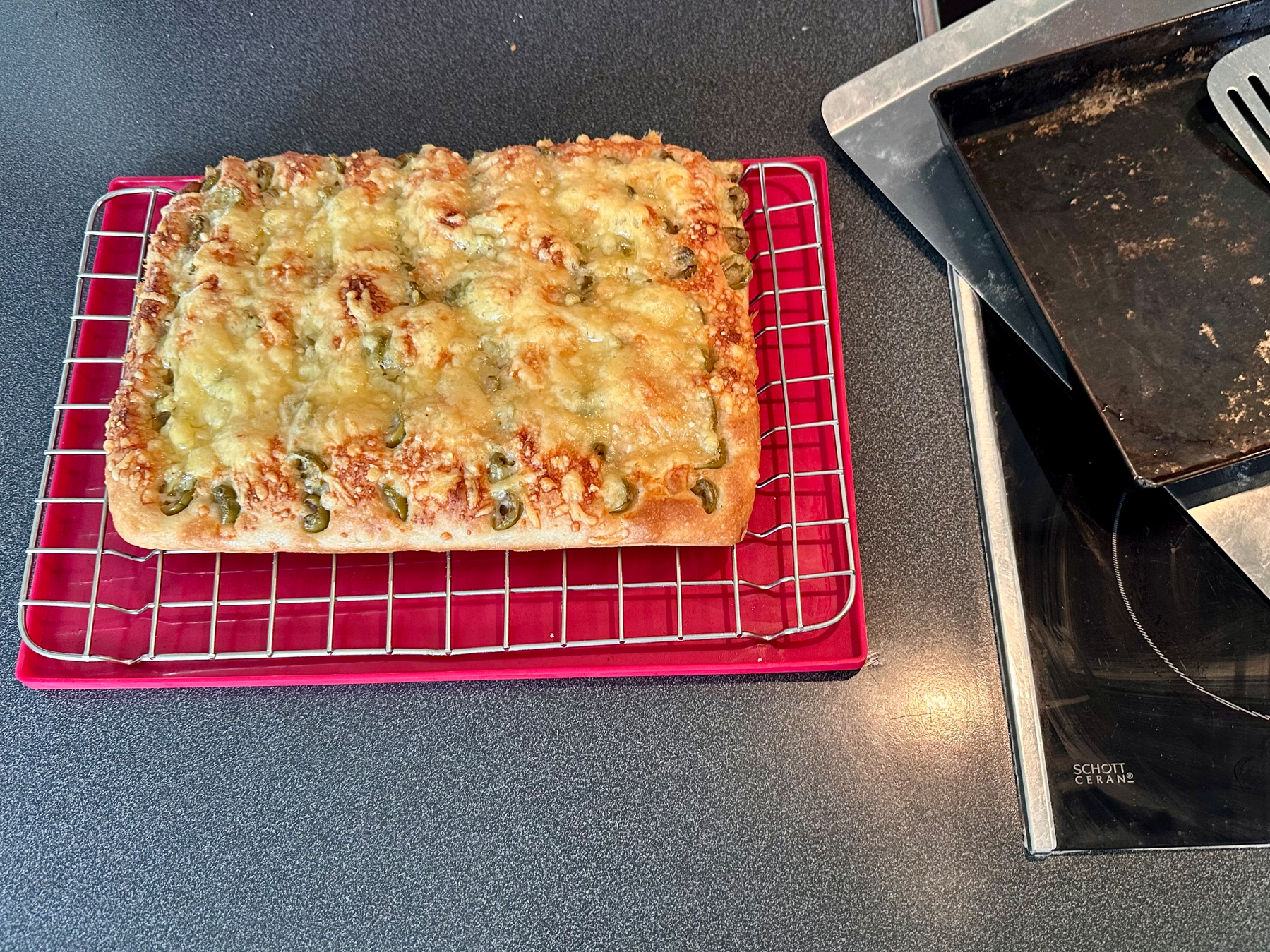 A freshly baked cheesy focaccia topped with herbs is resting on a cooling rack beside a stovetop and baking tray.