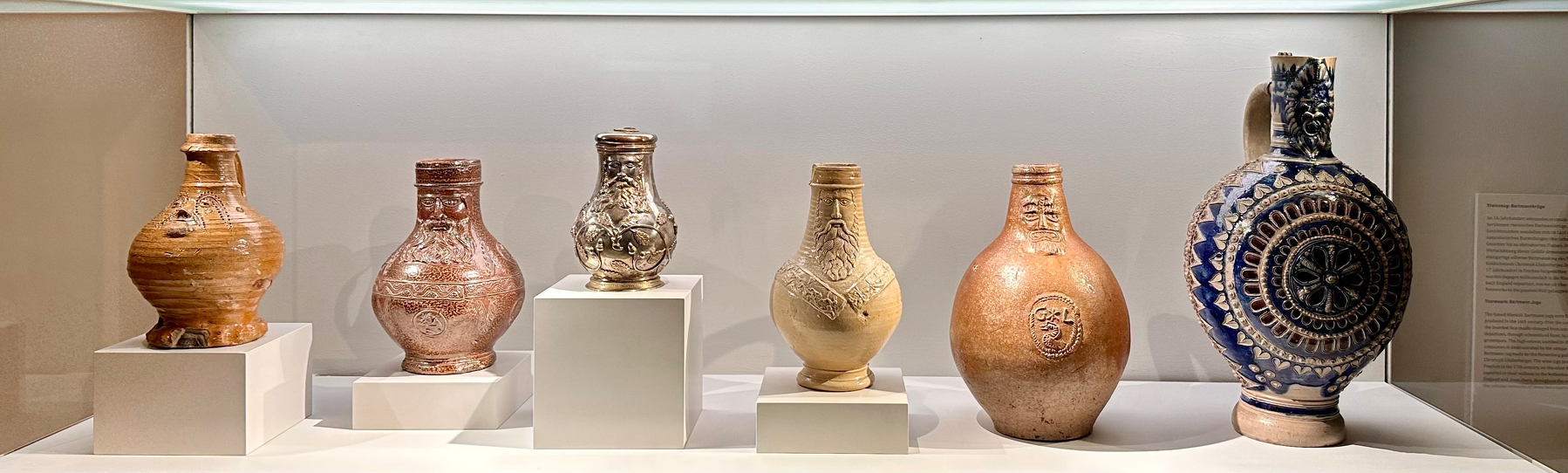 A display case features six vintage stoneware jugs of varying shapes and designs, each resting on white pedestals.