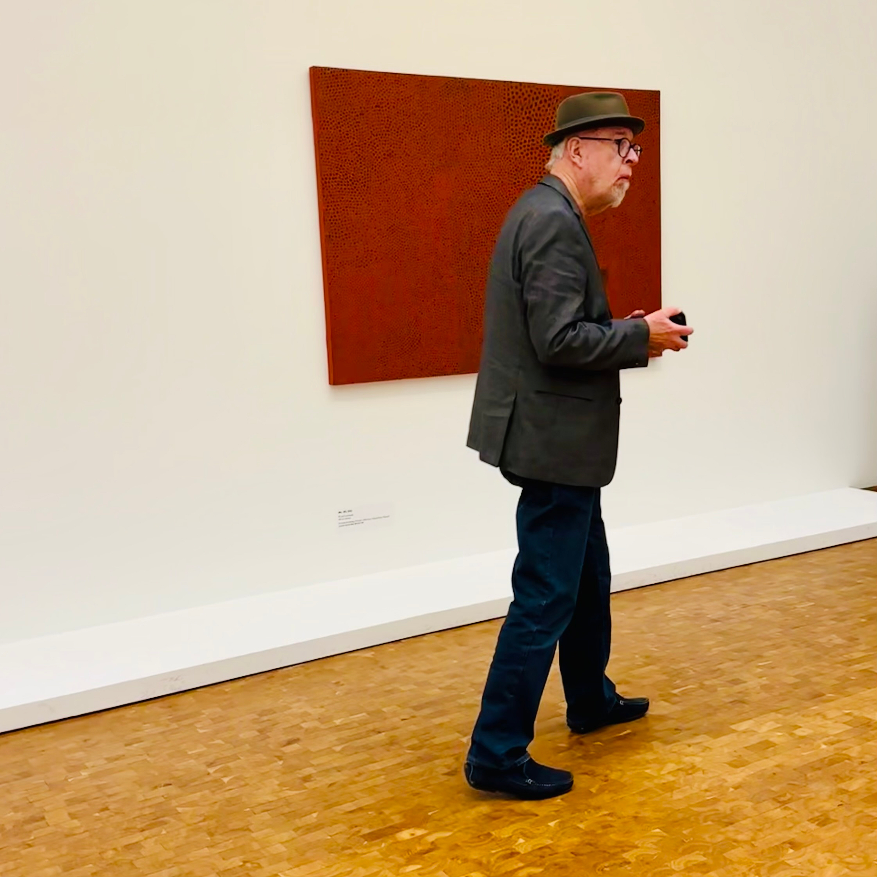 A man wearing a hat and glasses is walking across a wooden floor in an art gallery, observing a textured red artwork on the wall.