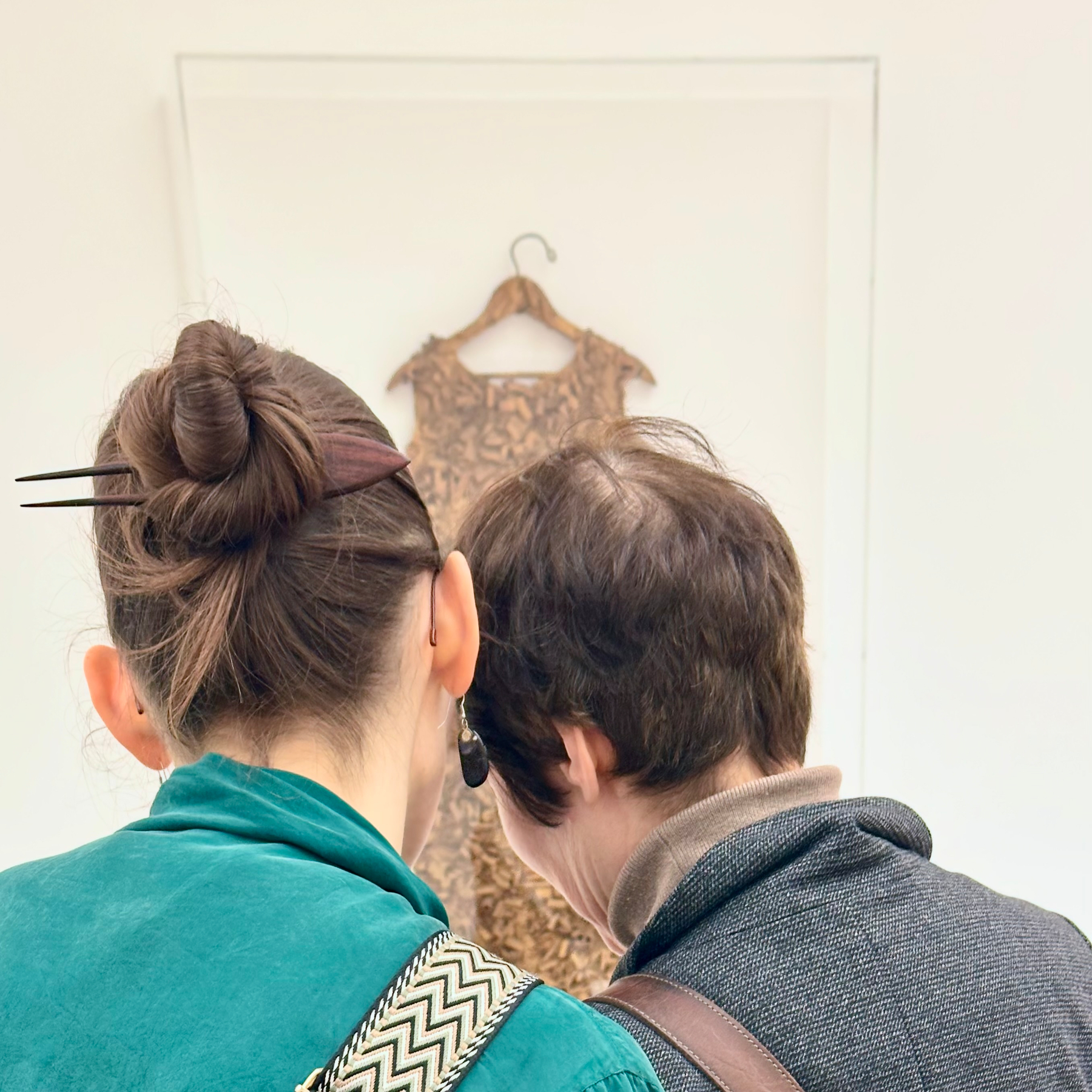 Two people are looking at a piece of art on a wall, with the person on the left having their hair styled in a bun with chopsticks.