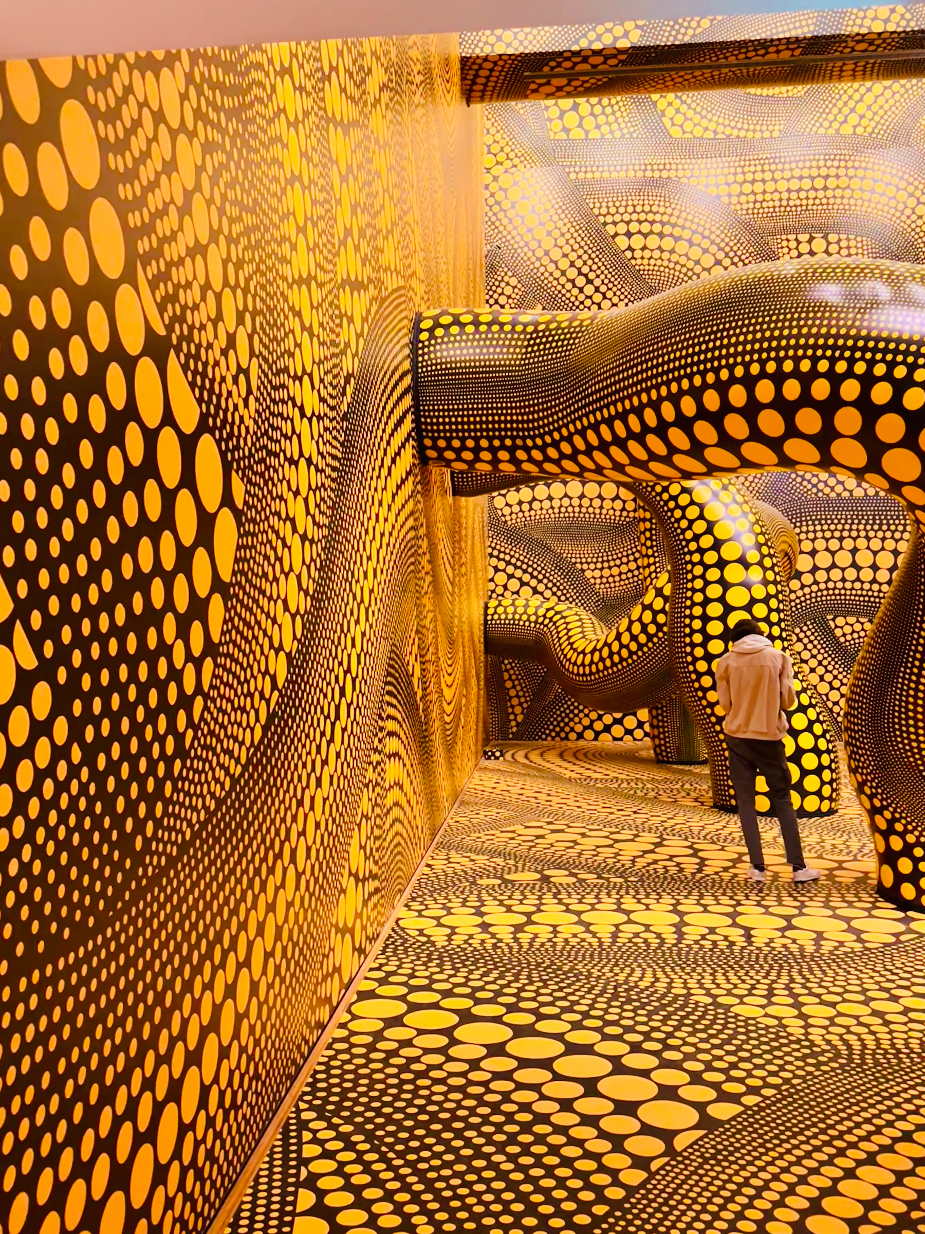 A person stands in a brightly colored, immersive room filled with intricate yellow and black polka dot patterns.
