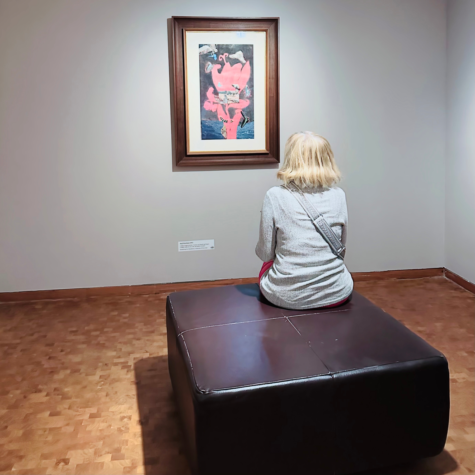 A person sits on a bench in an art gallery, viewing a framed painting on the wall.