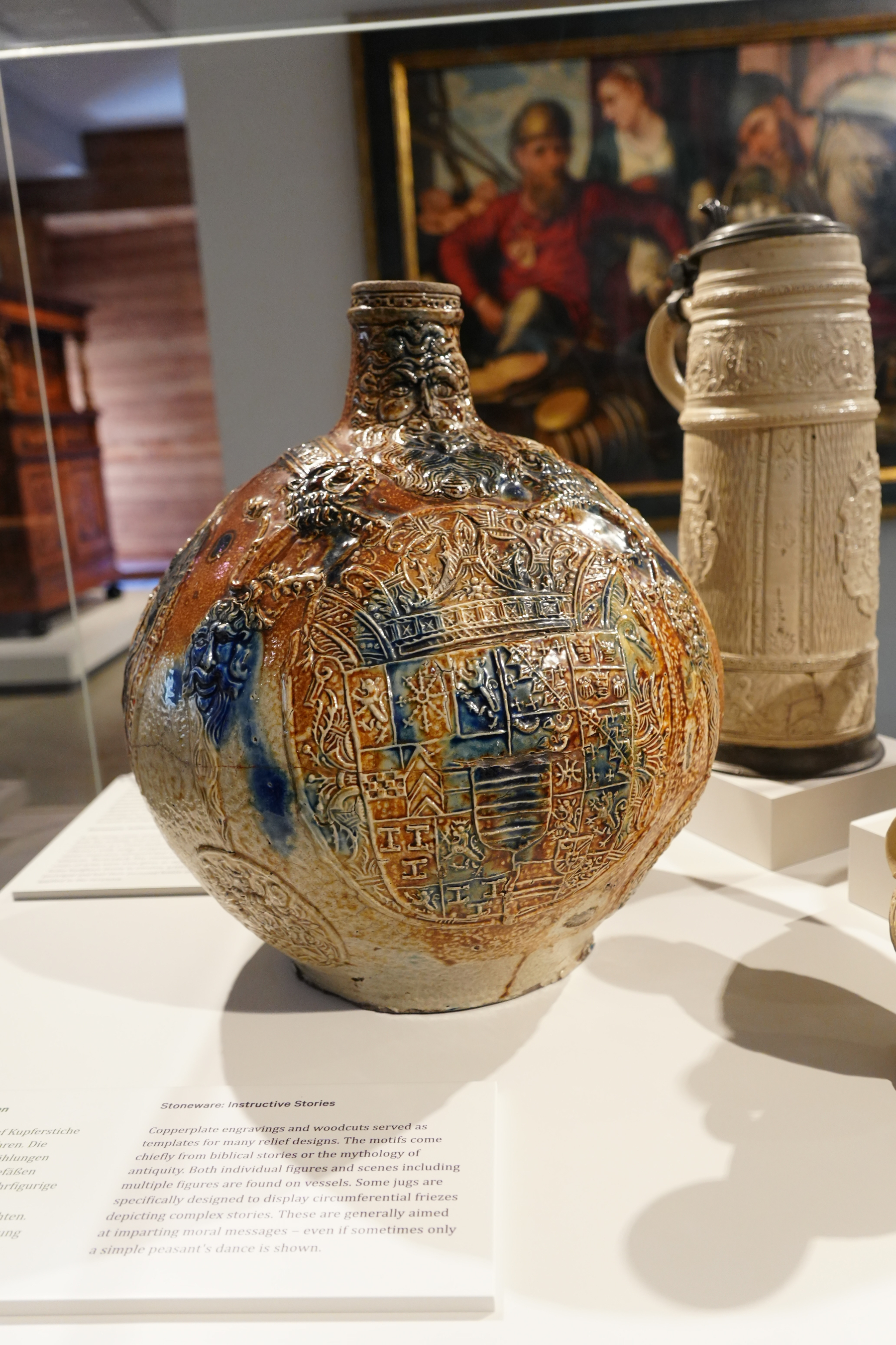 A large, ornate belarmine jug with intricate designs and a glazed finish is displayed in a museum setting.