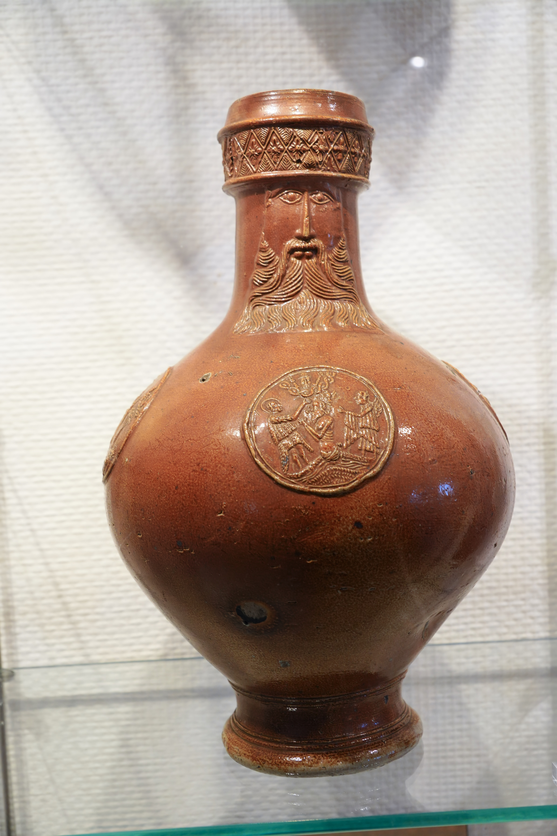 A brown, intricately designed jug features a bearded face and embossed medallions on its surface. A Bartmann Krug.