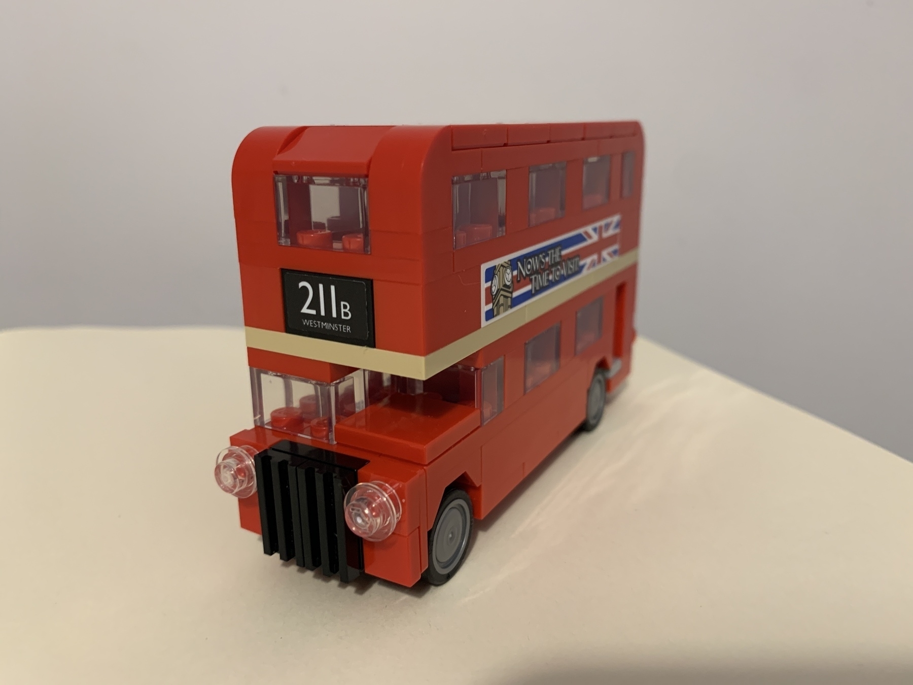 Small lego red London bus. The buses destination plate reads 211B Westminster. On the side of the bus is an adverstiment that reads Now is the time to visit. The background of the advert is the union flag and the top of Big Ben. The bus is sat on a cream colored base with a white/grey background
