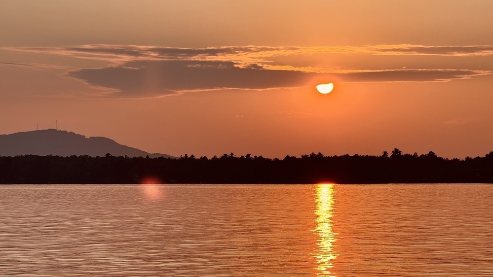 A serene sunset casts a warm glow over a tranquil lake with a silhouette of distant hills.