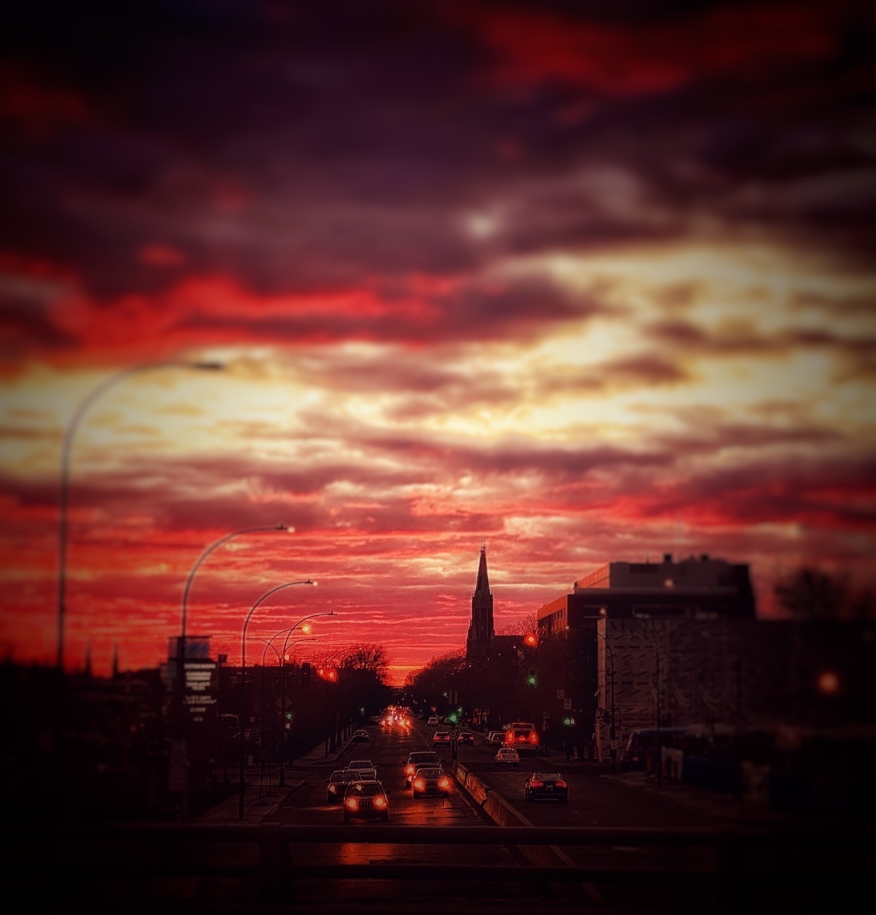 Auto-generated description: A dramatic sunset with vivid red and orange clouds looms over a city street, lined with cars and illuminated by streetlights.