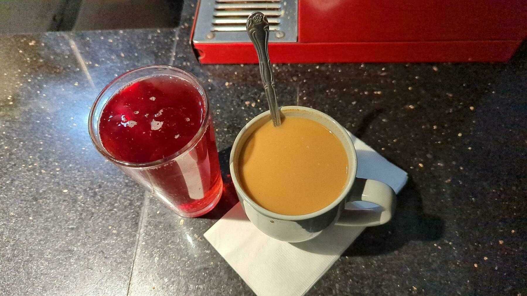 A wide white mug full of coffee with cream and sugar and a spoon sticking out of the top, next to a pint glass full of blackberry kombucha atop a dark marble countertop next to a bright red coffee grinder Sunday, March 26, 2023 at Rooz Cafe in the 1900 block of Park Boulevard in Oakland, California.