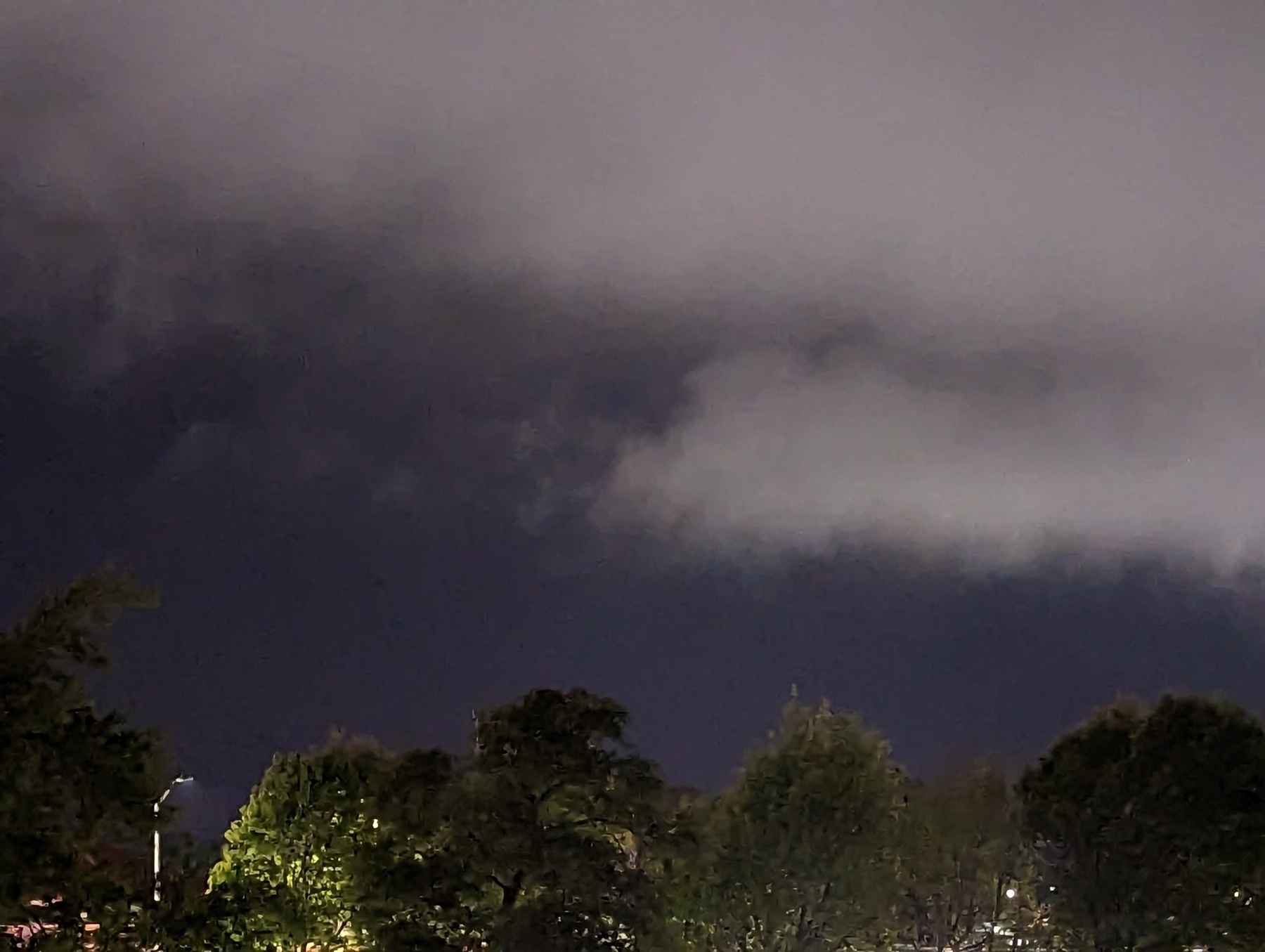 Pale gray puffy marine layer onshore flow clouds stream overhead, driven by winds from the south, against nearly navy blue night sky over the lamp illuminated tree topped western skyline Friday April 28, 2023 in San Pablo, California.