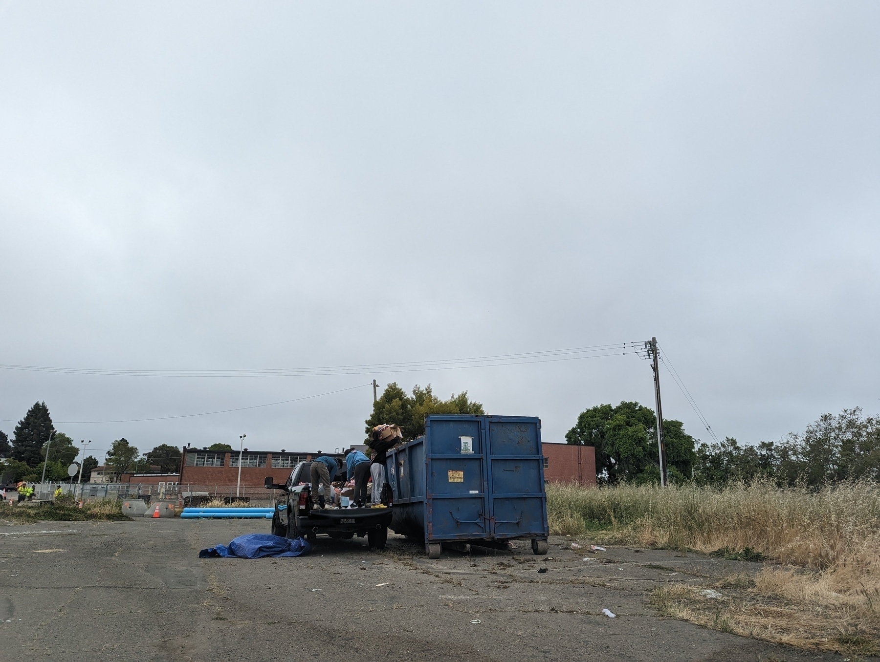 A pair of people drop off refuse and other items from the rear bed of a pickup truck into a big blue dumpster in a wide open and empty but weed choked lot under low light gray cloudy skies Saturday, May 20th, 2023 at the San Pablo public works department in the 2600 block of Moraga Road in San Pablo, California.