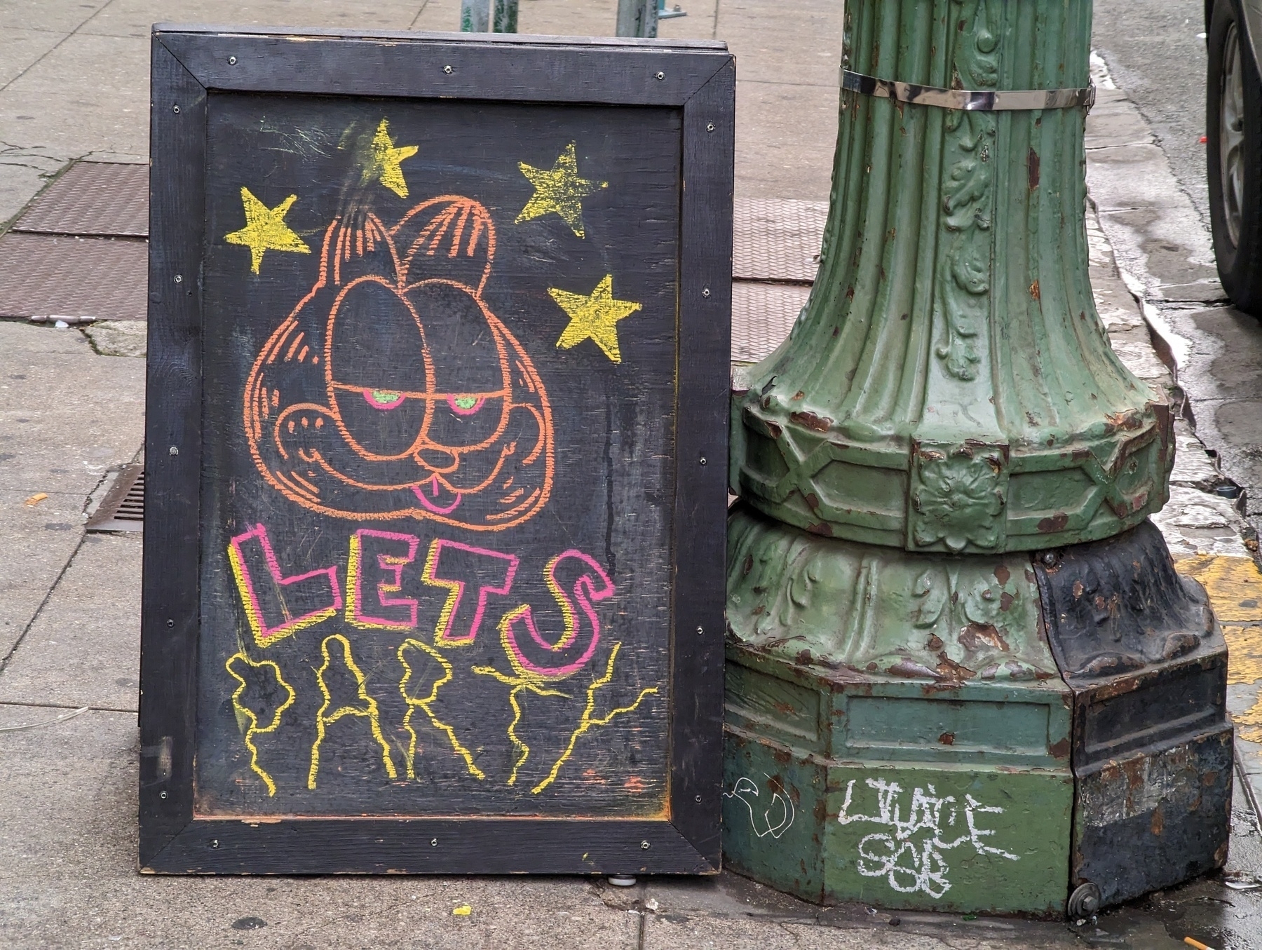A chalkboard sandwich-board sign with a colored chalk caricature of Garfield the cat on it surrounded by solid yellow stars over the scribbled words 'let's party' rests on a sidewalk next to a green painted base of a street lamppost Sunday, March 12, 2023 in the 400 block of 14th Street in Oakland, California 