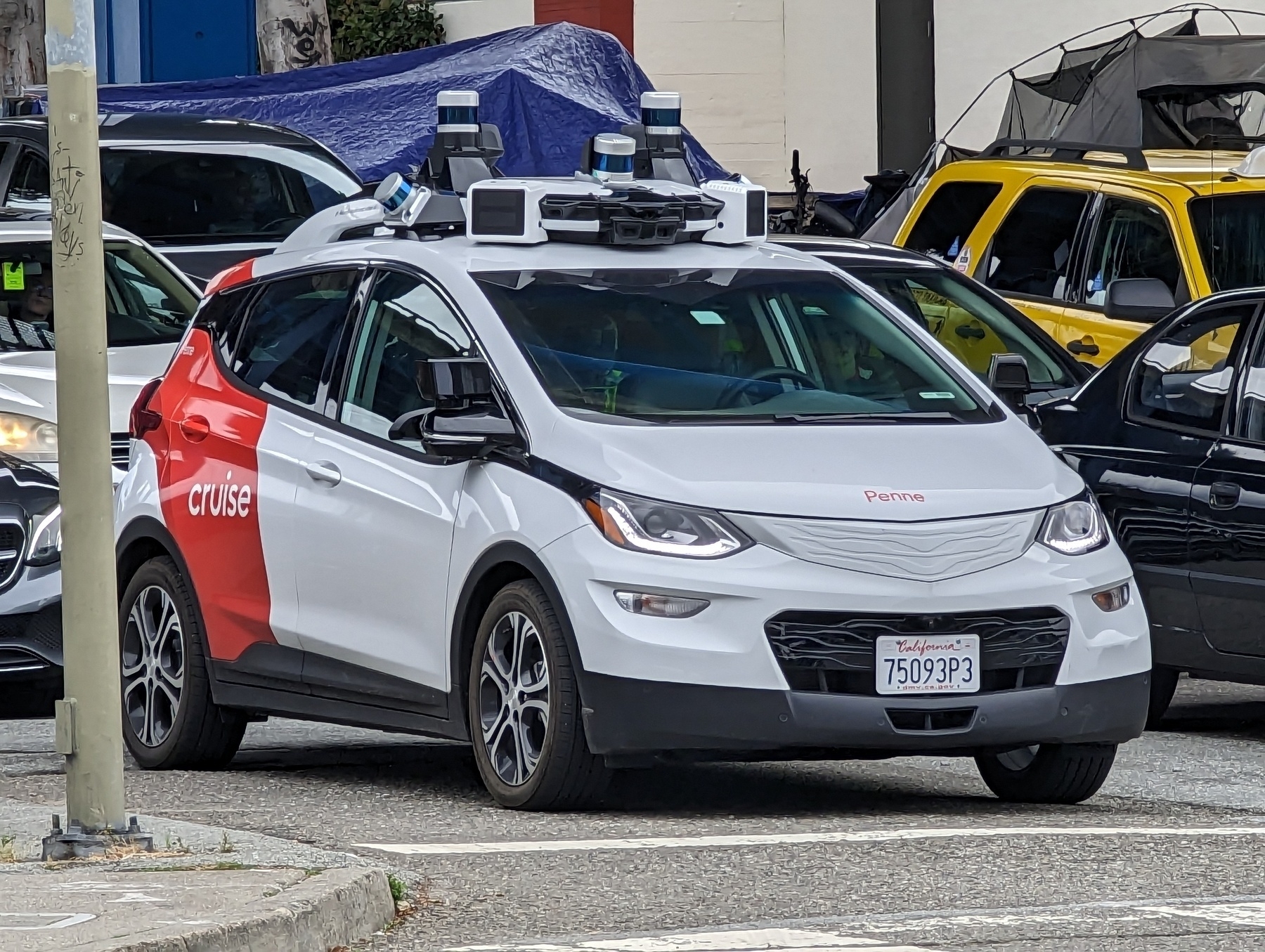 A driverless Cruise car, a compact white four door sedan with a roof almost completely covered in cameras and mounted sensors, waits for a green light Friday, May 5, 2023 in northbound Portrero Avenue lanes at Division Street in San Francisco, California. On sidewalks behind and beyond the car, blue plastic sheeting and thin metal poles tied to gray nylon fabric show separate tents unsheltered city residents have set up.
