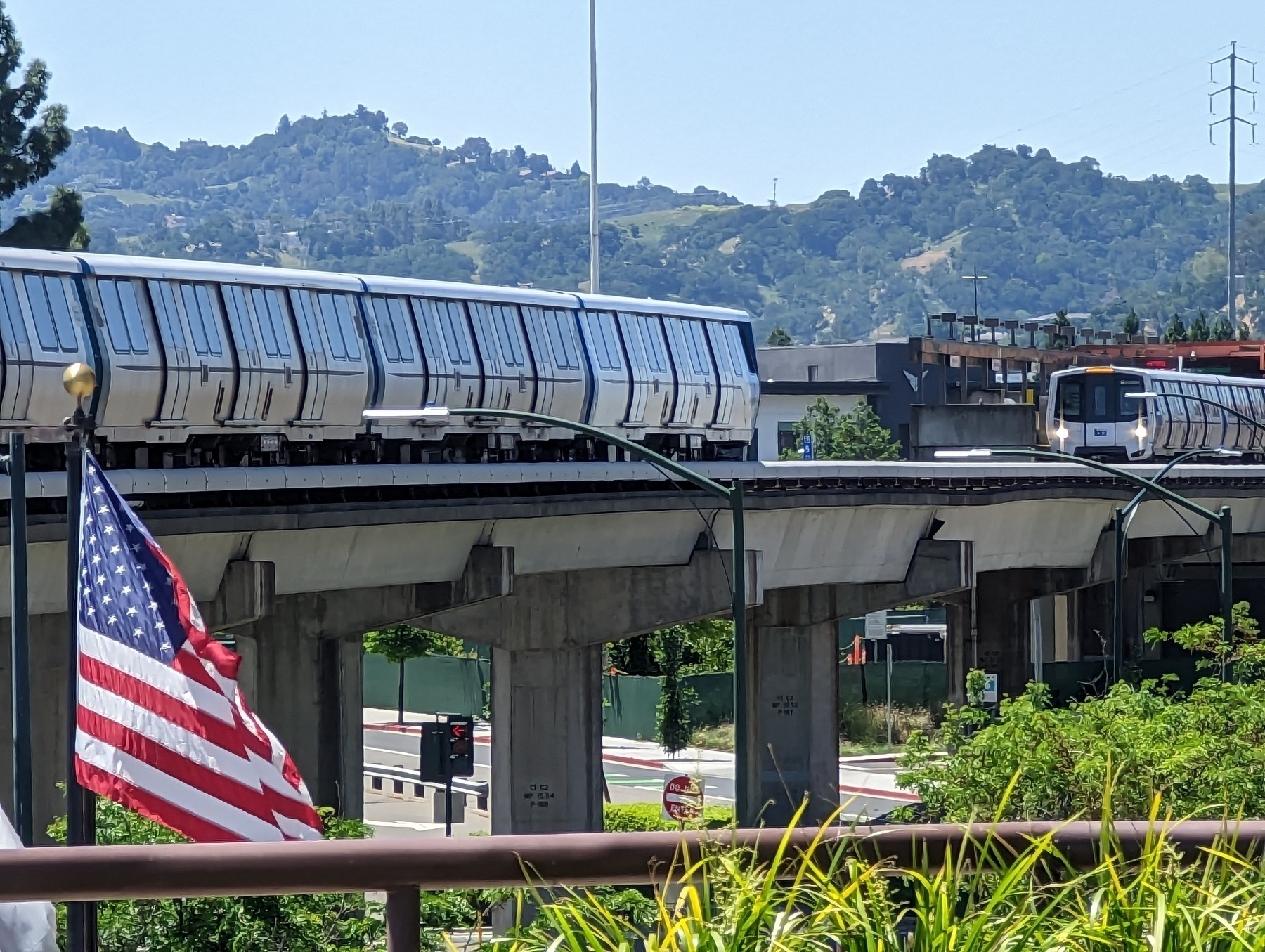 A view of an American flag beside an elevated BART track with two trains running along it next to the Walnut Creek BART station as seen from the California Plaza office complex Wednesday, May 17th 2023 in the 2100 block of North California Boulevard in Walnut Creek, California