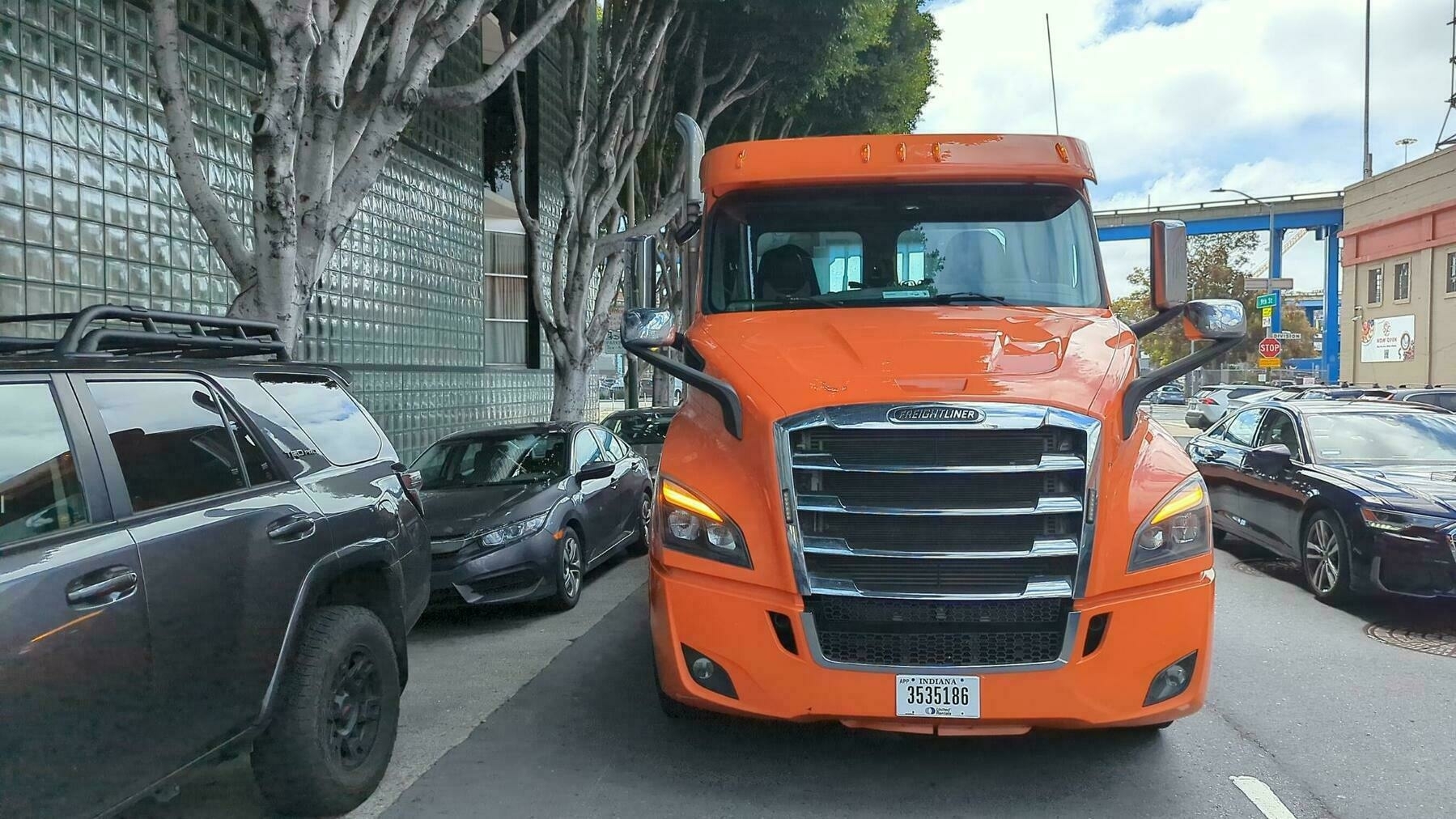 A bright orange painted flatbed Freightliner truck with Indiana plates sits in traffic beside a row of parked vehicles and tall well manicured trees and a building with a large glass bricked first floor lobby Thursday May 12th 2023 on Ninth Street in San Francisco, California.