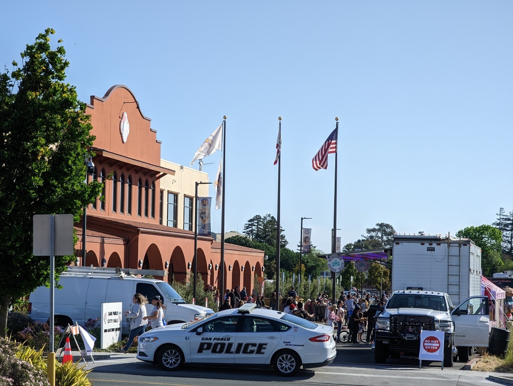 A white San Pablo Police Department patrol vehicle blocks off the entrance to the long driveway Thursday, April 27th 2023 at the 75th anniversary celebration outside City Hall in San Pablo, California. Above a small crowd enjoying food trucks and lowrider classic cars, three flag poles show the American, state and city flags against clear pale blue afternoon sky.