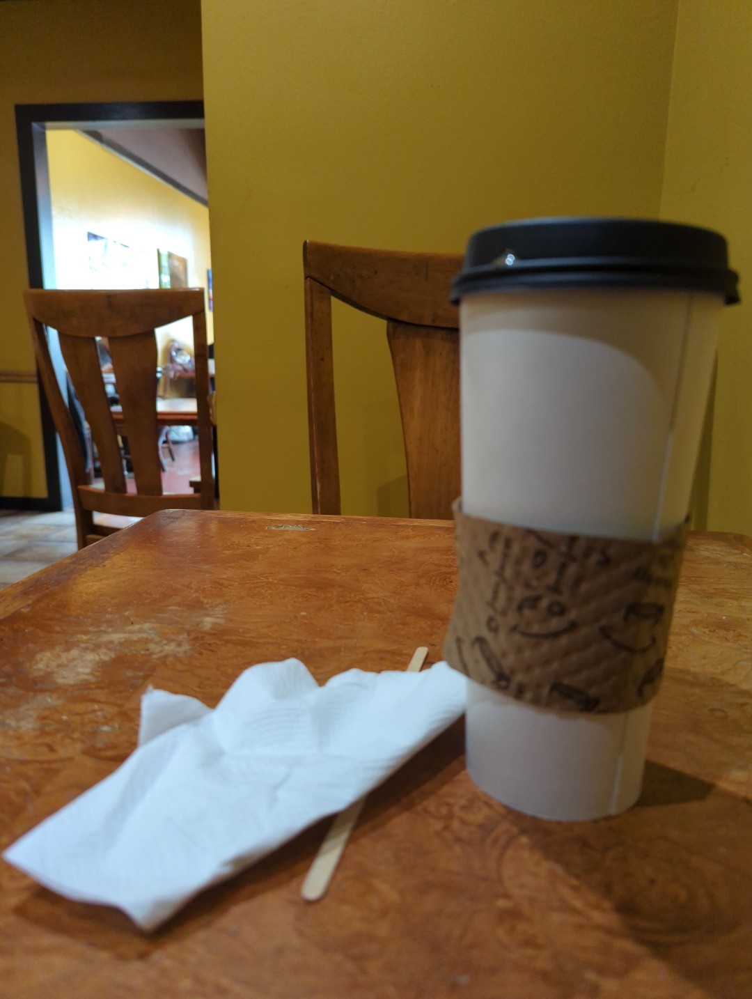 A slightly out of focus picture of a 24 ounce white paper coffee cup next to a napkin and coffee stirrer on a wooden surface table with two wooden chairs nearby in a back aisle of World Grounds Cafe Sunday around 10 a.m. April 23, 2023 in Oakland California's Laurel district.