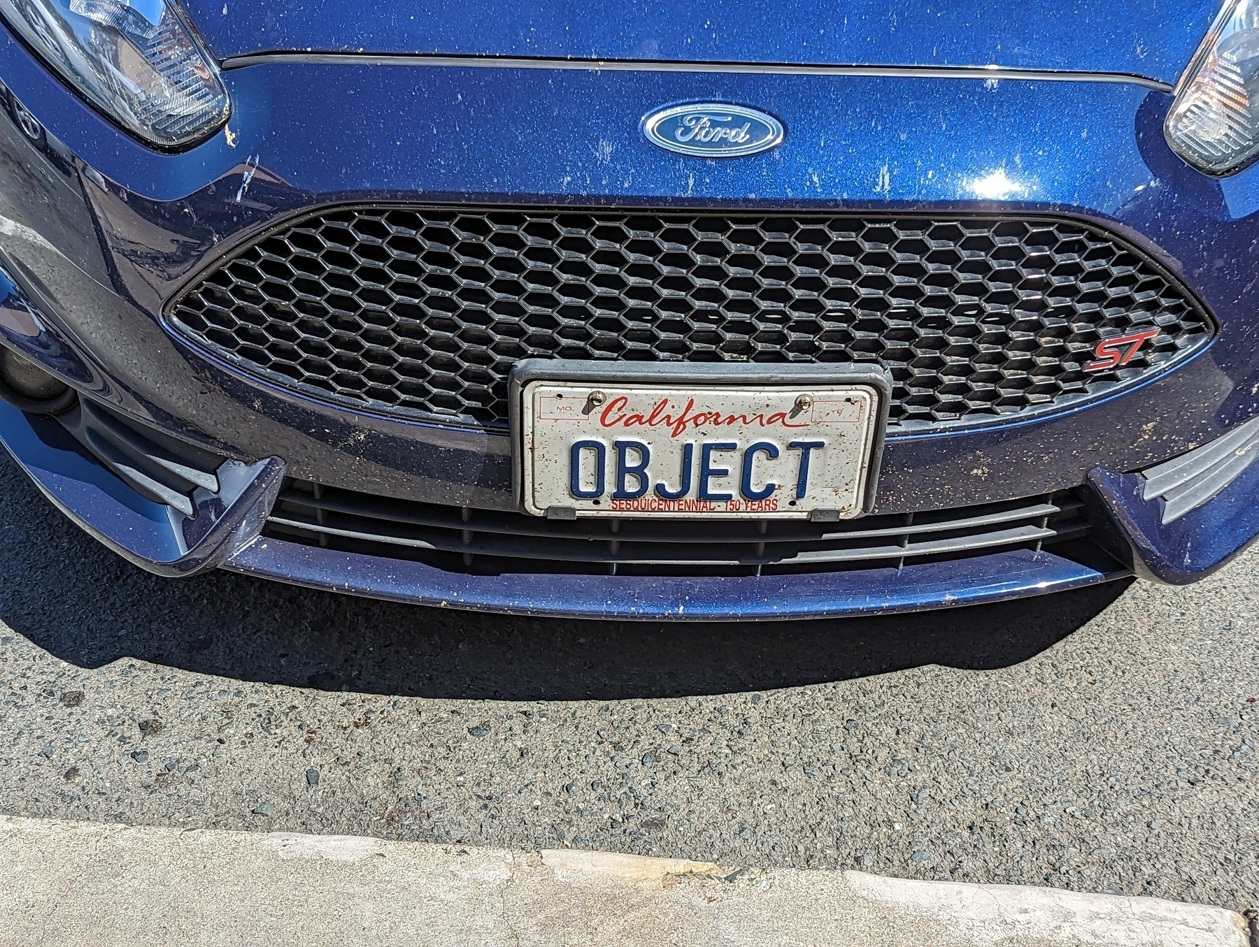 A custom California license plate with the word 'OBJECT' sits on the front end of a blue Ford sedan in the parking lot outside a Whole Foods Market store Saturday, May 13, 2023 in Walnut Creek, California.