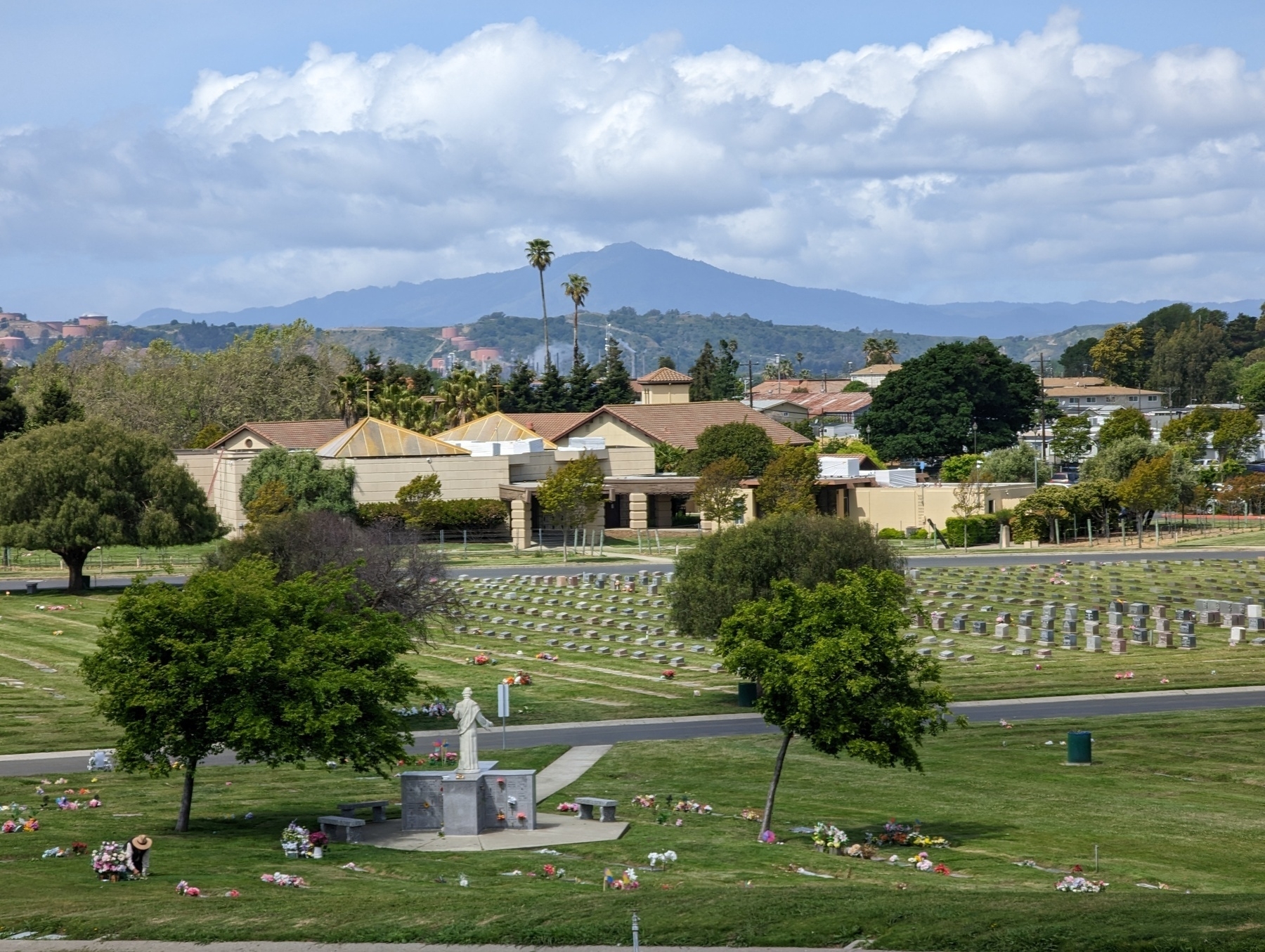 A zoomed in (4x magnification) view facing west toward Mount Tamalpais from the top of a roadway winding through St. Joseph's Catholic cemetery on Church Road with rows of flower and flag adorned gravestones under partly sunny skies Tuesday May 9, 2023 in San Pablo, California.